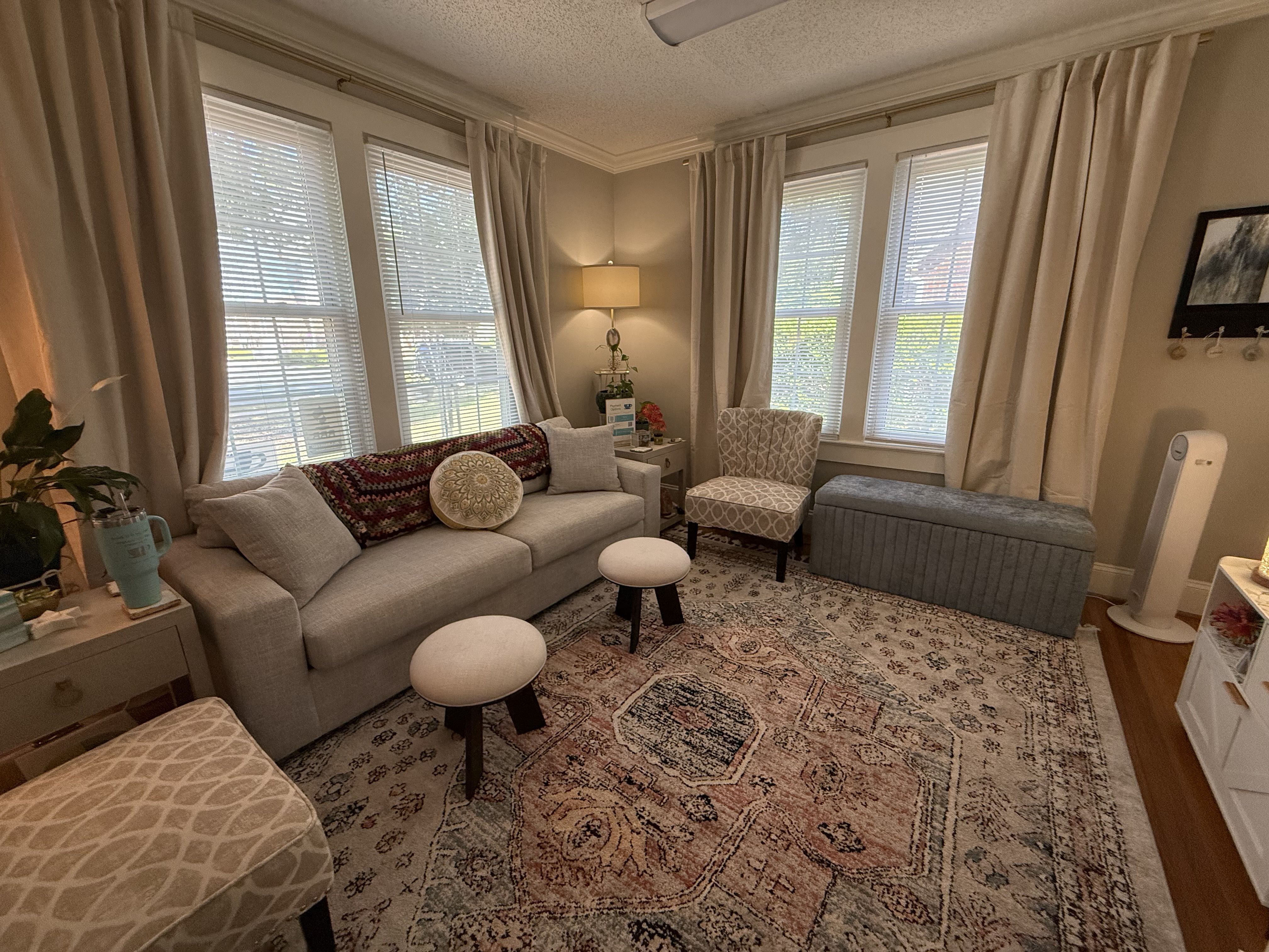 Cozy living room with beige walls and tall windows with blinds and cream curtains. A beige sofa with a patterned throw, round pillows, ottomans, a patterned rug, and a lamp on a side table.