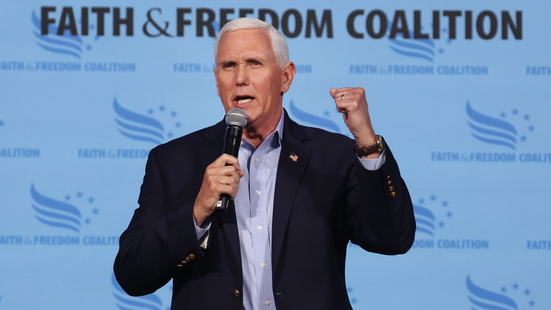 Mike Pence stands in front of a blue banner reading 'Faith and Freedom Coalition' while holding a microphone as he speaks to a crowd.