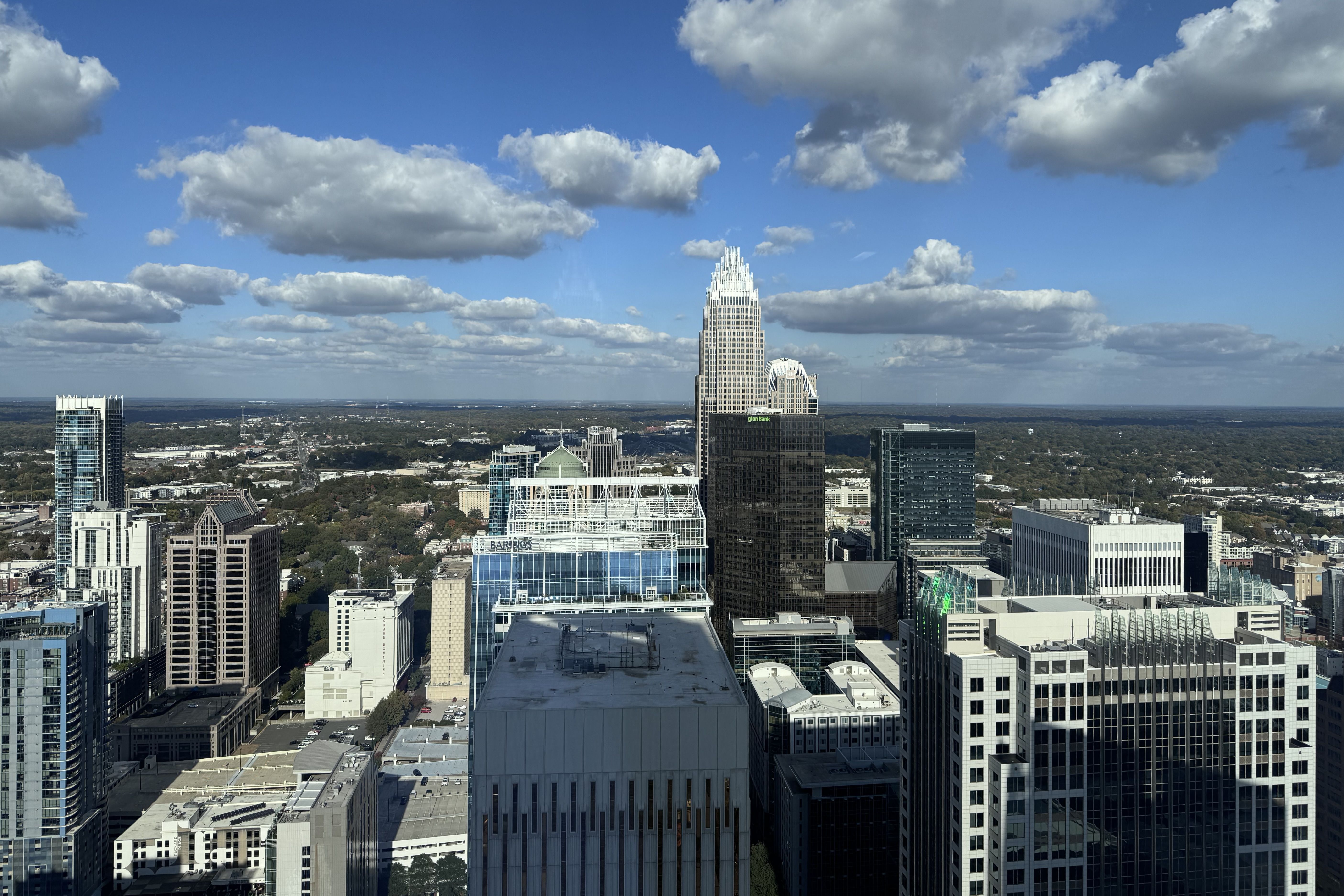 City skyline under a blue sky with scattered white clouds. A central white, stepped, pyramid-like skyscraper rises above surrounding glass office towers.