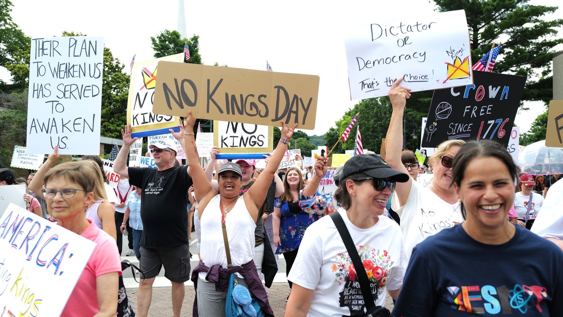 Smiling protesters march with signs like "NO KINGS DAY" and "Dictator or Democracy," waving flags and calling for democracy.