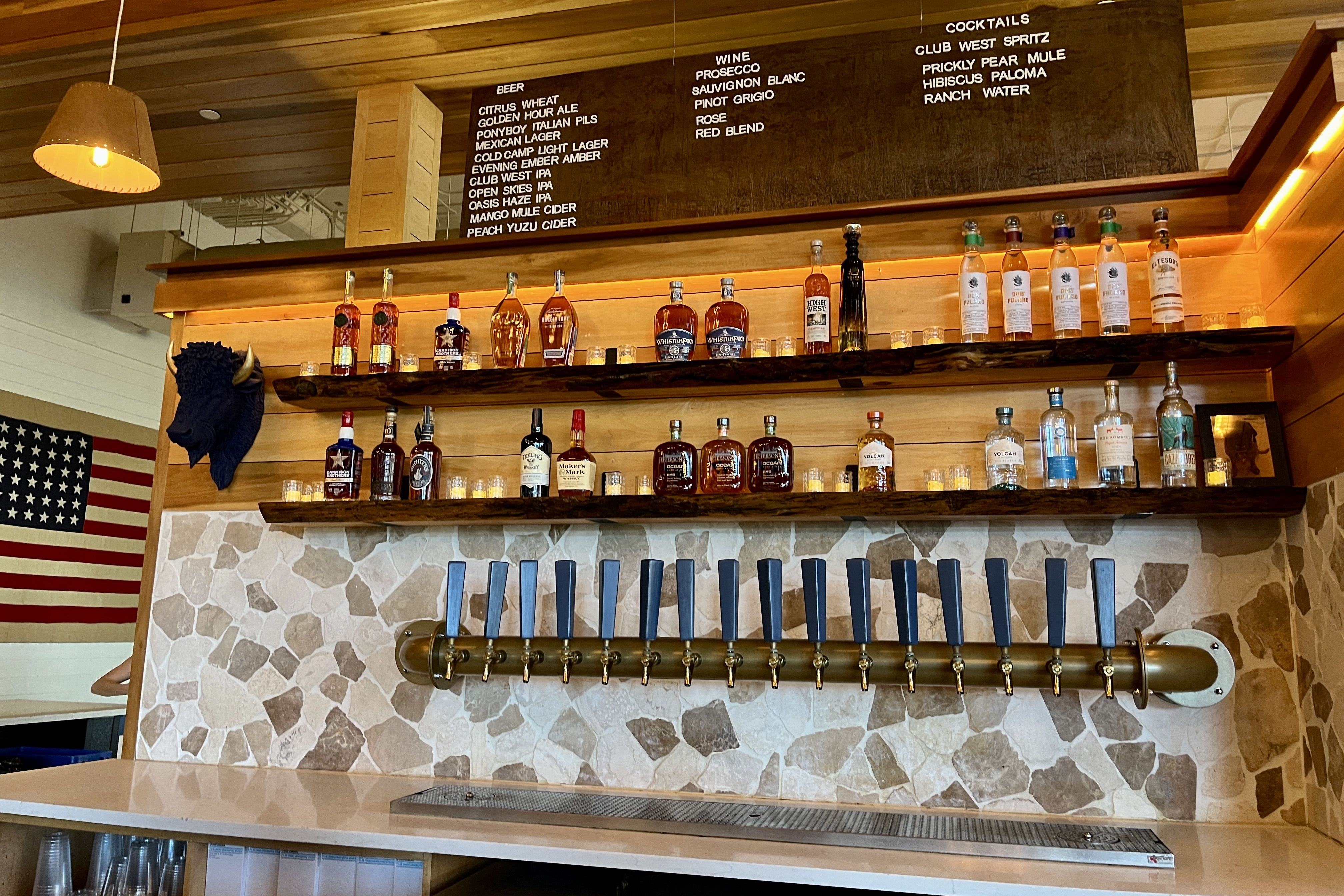 Bar interior with a long row of blue beer-tap handles on a brass rail, wooden liquor shelves, a stone backsplash, an American flag, and a black mounted animal head.