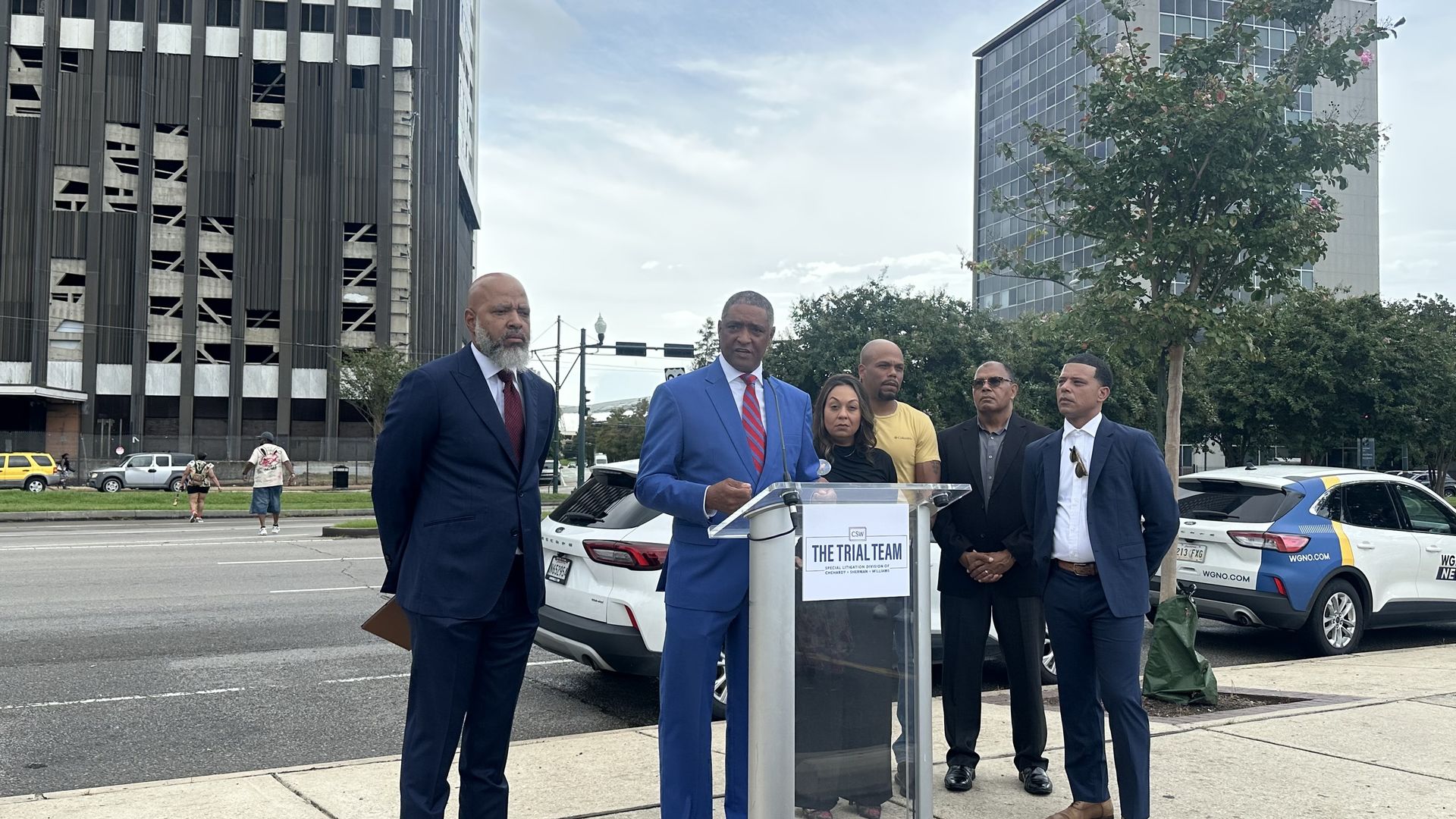 Group of professionals at a podium labeled "The Trial Team" on a city street; a man in a blue suit speaks, flanked by other people, with cars and Plaza Tower in the background.