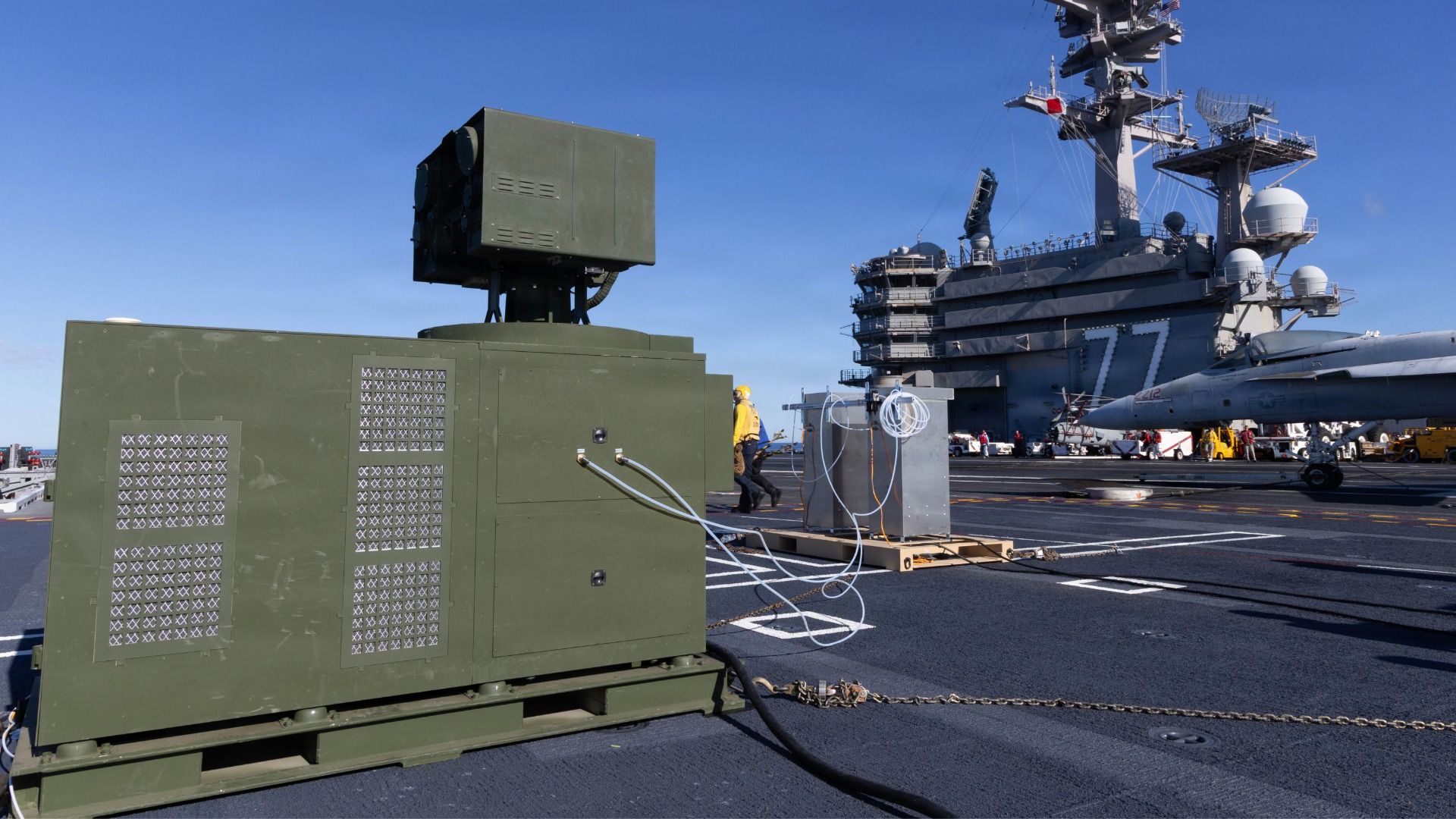 A green-in-color laser weapon on a Navy aircraft carrier deck. The sky overhead is clear and blue.