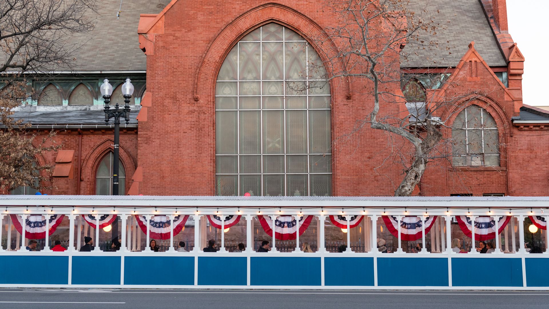 Red brick Gothic-style building with a tall arched window, leafless trees, and a vintage lamppost in front. A streatery in front with red, white, and blue bunting and blue panels runs along the base.