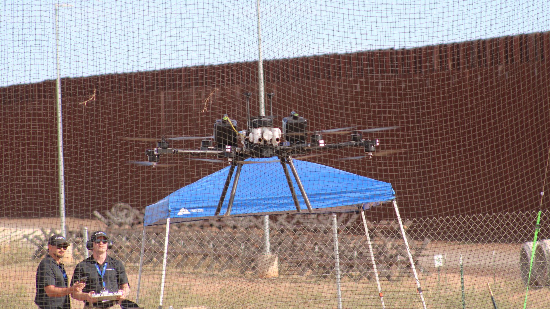 A large, black, multi-rotor drone hovers in front of a blue awning in a netted area in the desert, with two technicians controlling it and a tall, copper-colored steel fence in the background. 