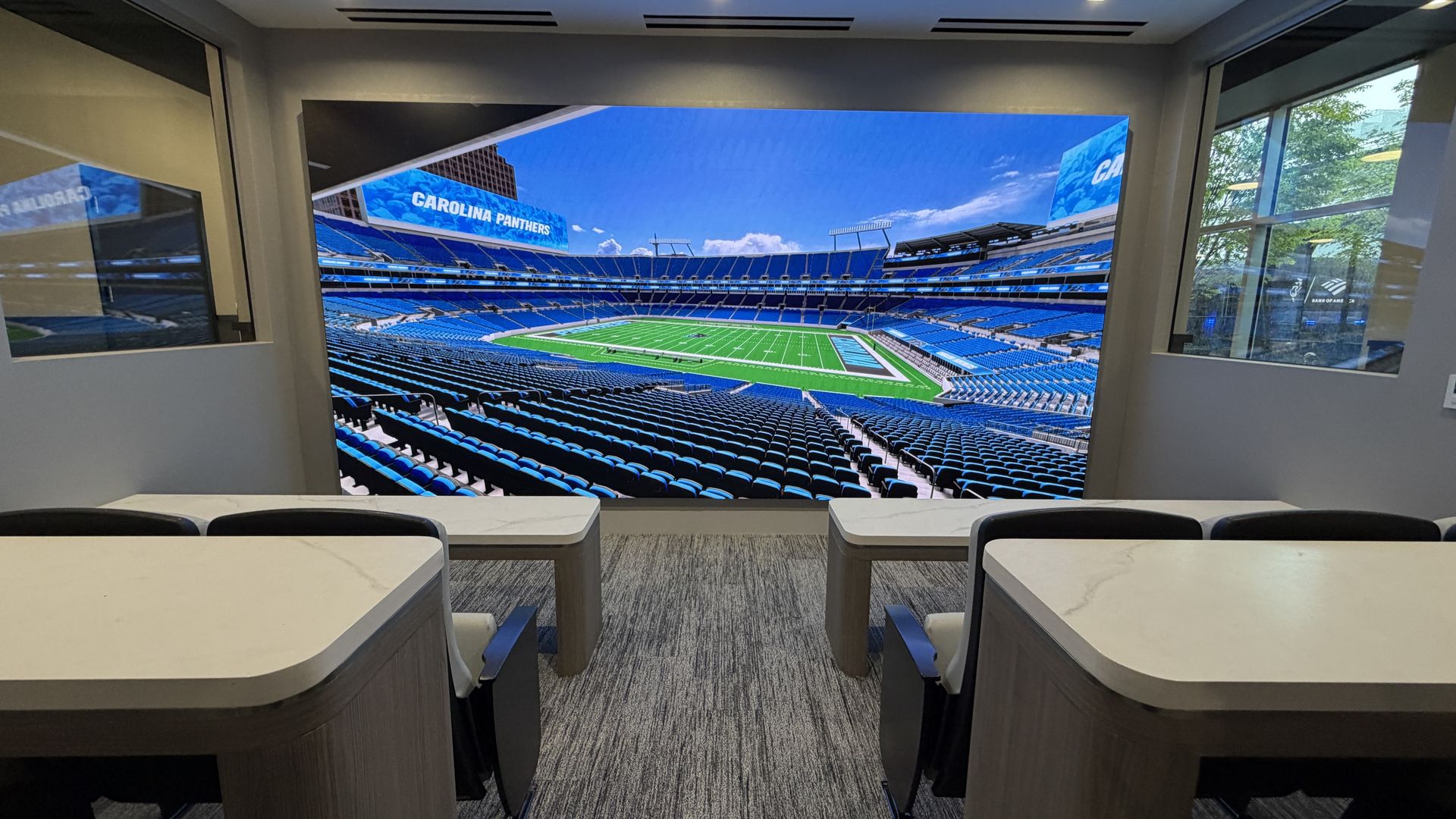 Stadium suite with a large wall screen showing the Carolina Panthers stadium: blue seats surround a green field; empty stands, desks in the foreground.