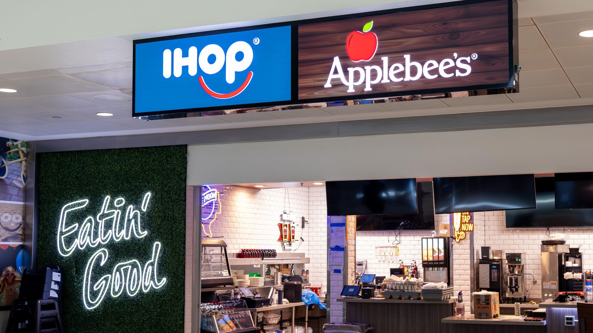 Interior of a dual restaurant at Dallas Love Field featuring illuminated IHOP and Applebee's signs above a kitchen area, with a neon sign on the left reading "Eatin' Good" on a green backdrop.