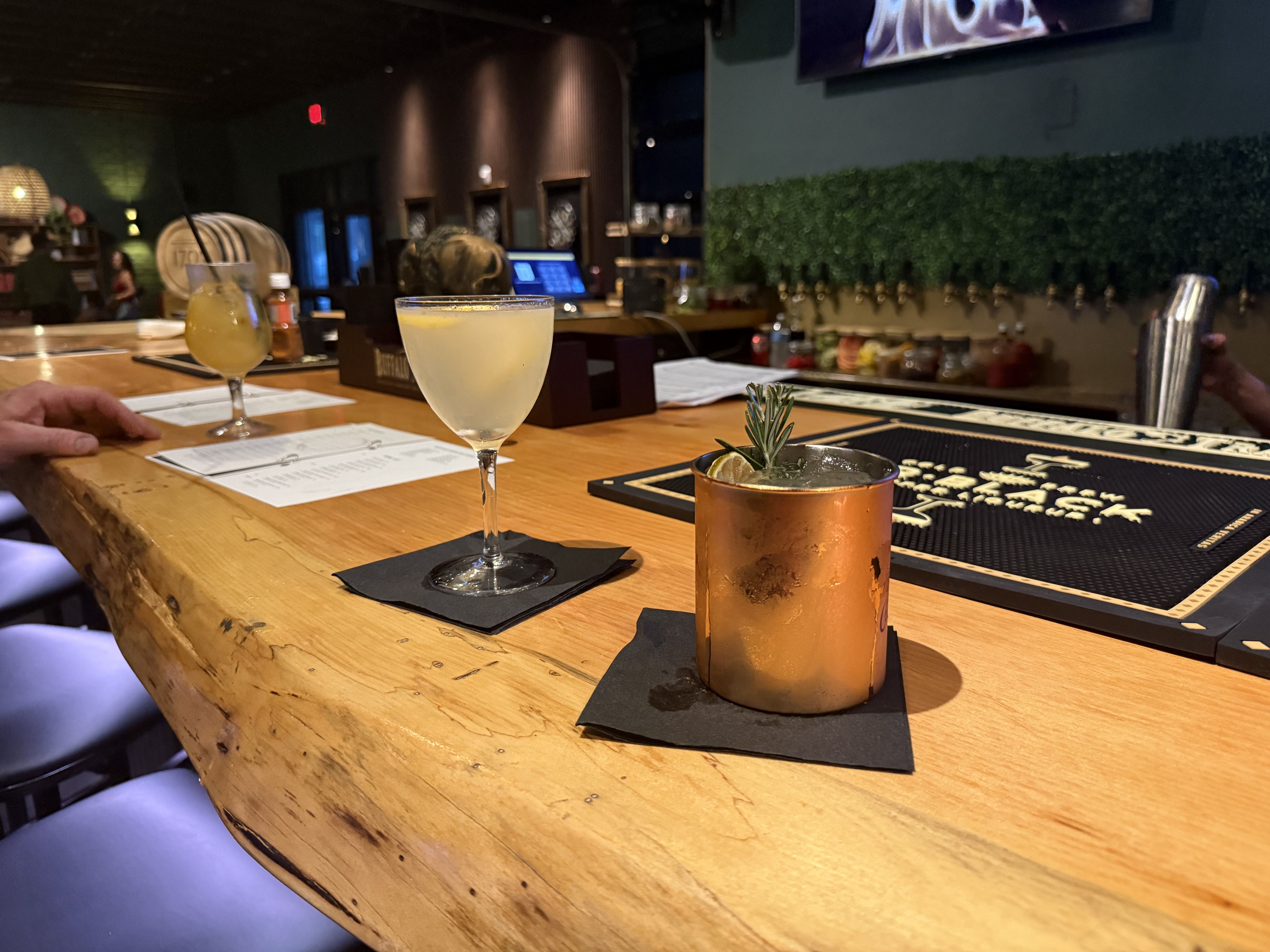 Dim bar interior with a wood counter. In the foreground a pale yellow cocktail in a wine glass and a copper mule mug with lime. Background shows shelves, a green hedge wall, and a blurred bartender.
