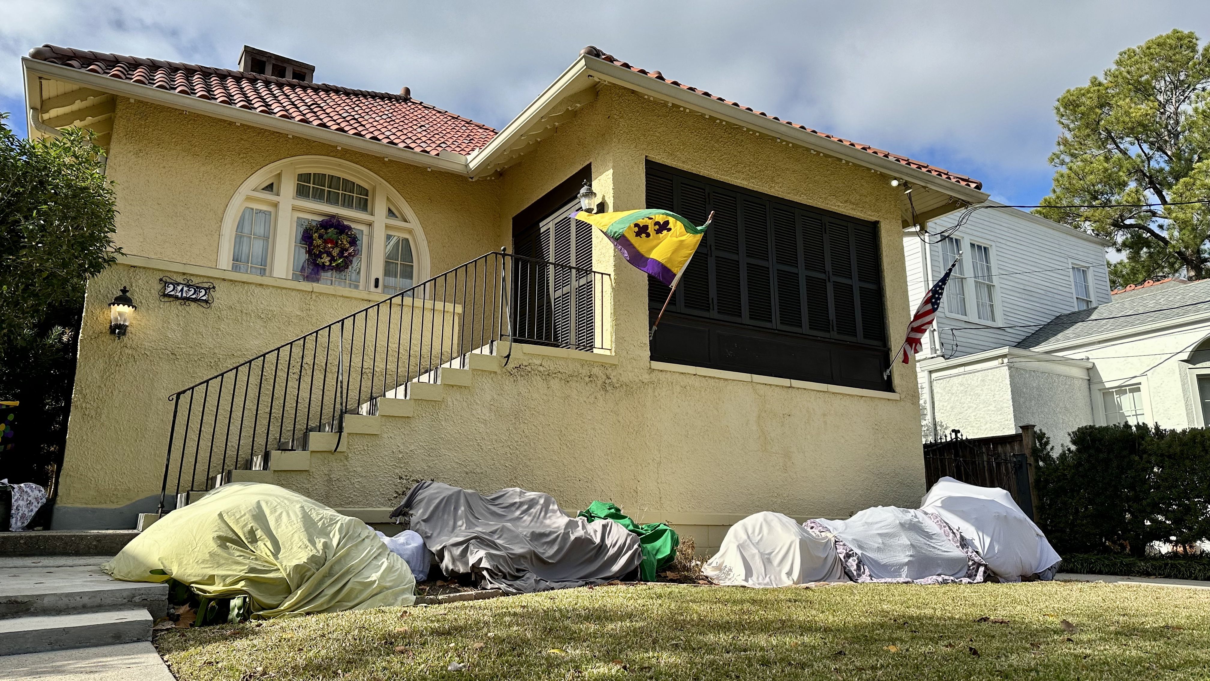 Photo shows a house with a Mardi Gras flag and covered plants.