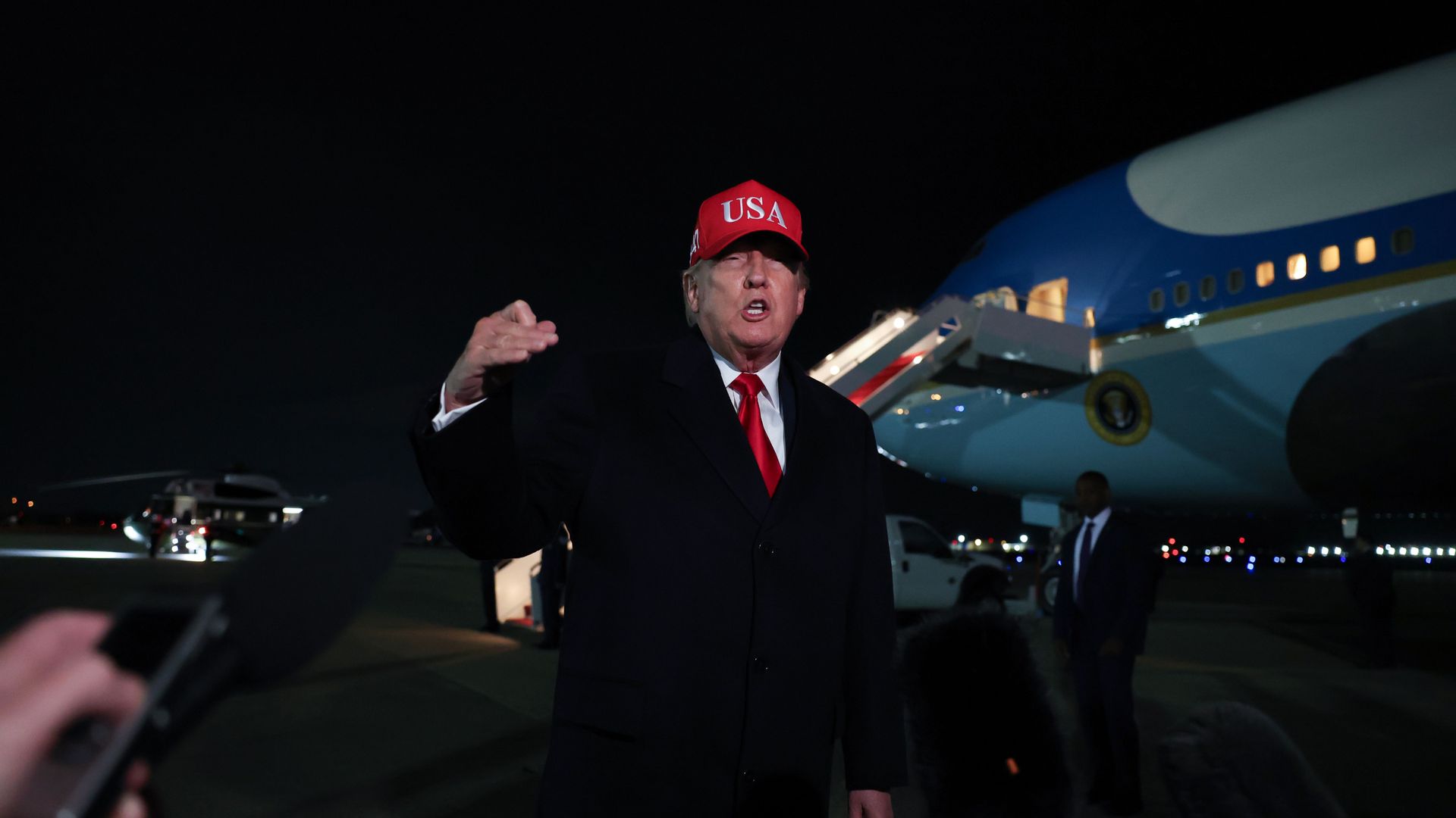A man in a dark suit and red tie, wearing a red cap with USA, addresses reporters on a nighttime airport tarmac as a large blue-and-white jet looms behind him; microphones visible in the foreground.