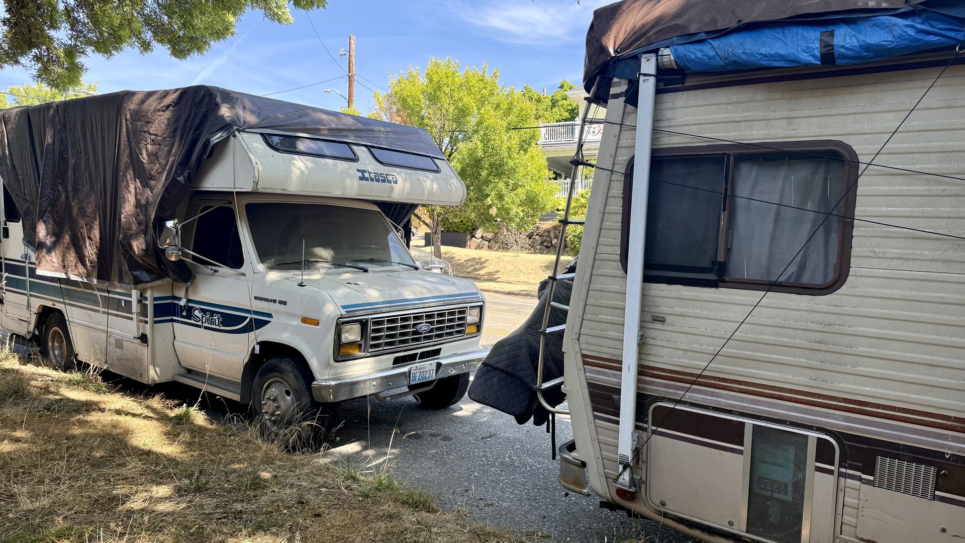 Two parked RVs under a tree on a sunny day: a white motorhome with teal stripes on the left, covered by a dark tarp, and a beige trailer on the right with a blue tarp.