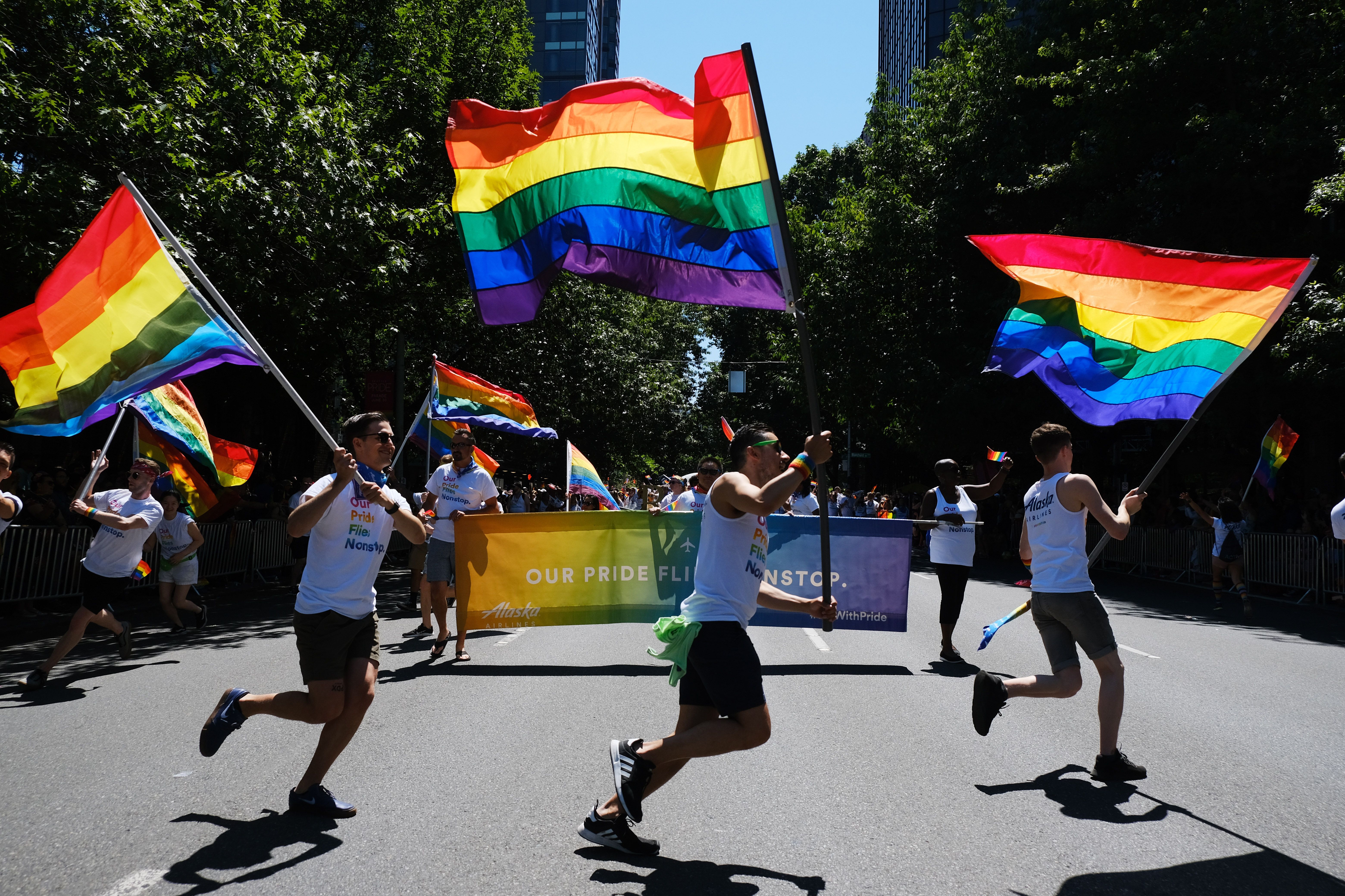 Three people in the foreground during a parade march while carrying rainbow Pride flags.