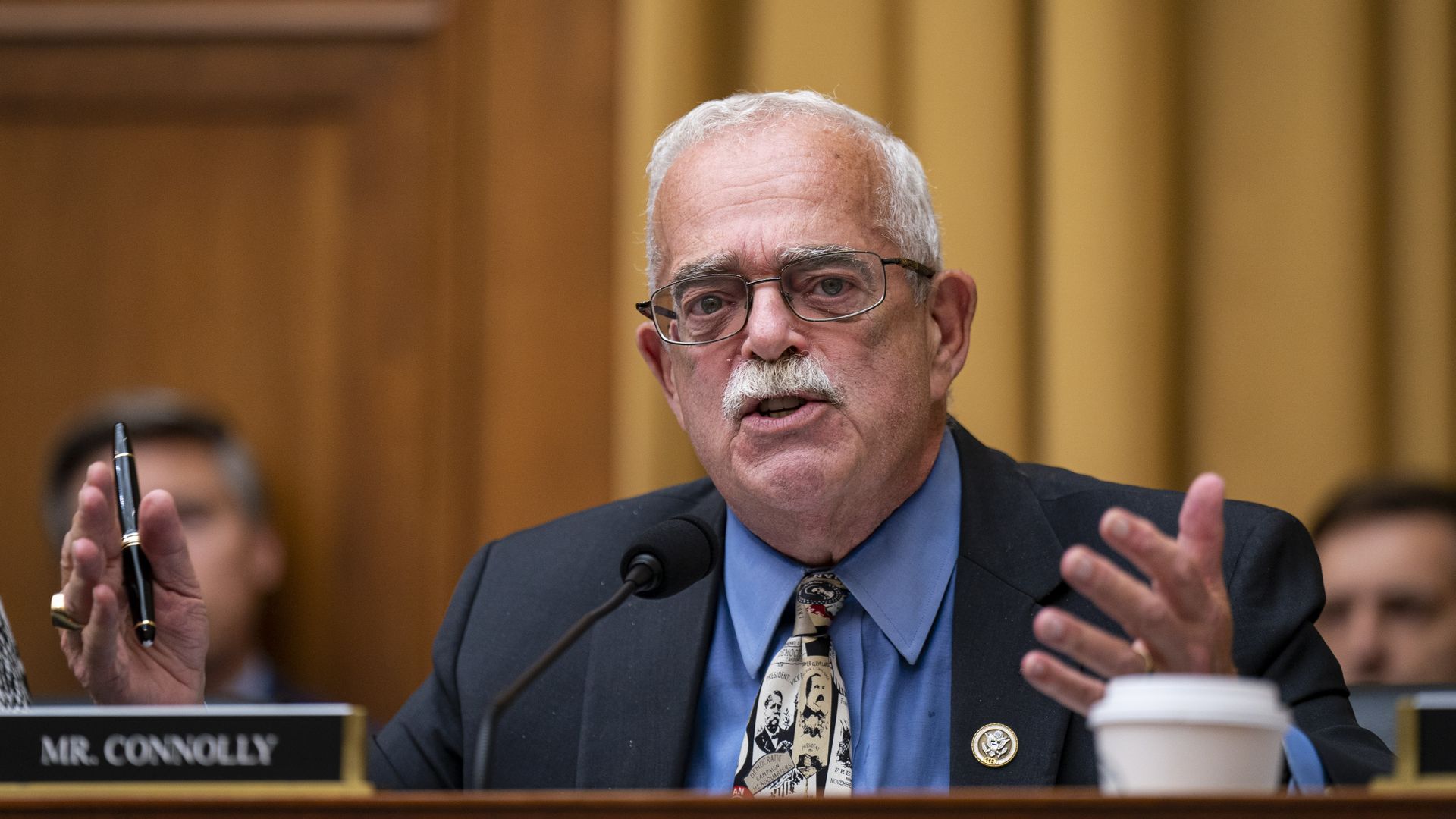 Rep. Gerry Connolly, wearing a gray suit, blue shirt and yellow tie and speaking into a microphone at a committee dais. 