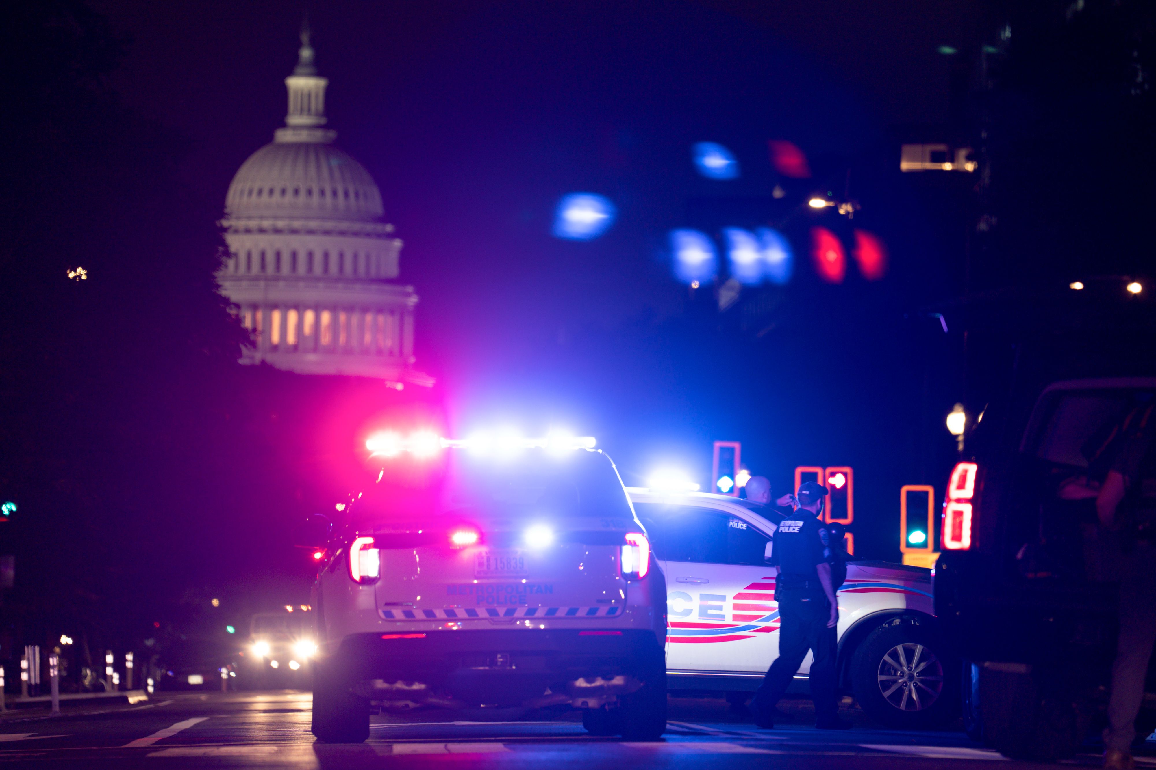 Two police cars with flashing red and blue lights at night near the illuminated U.S. Capitol building dome. Police officers stand beside the vehicles on a dark street.