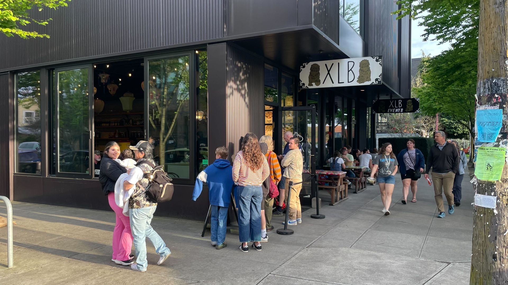 People wait in line outside XLB, a modern Asian restaurant. Others walk by or sit at outdoor tables. It's a sunny day with green trees and flyers posted on a utility pole nearby.