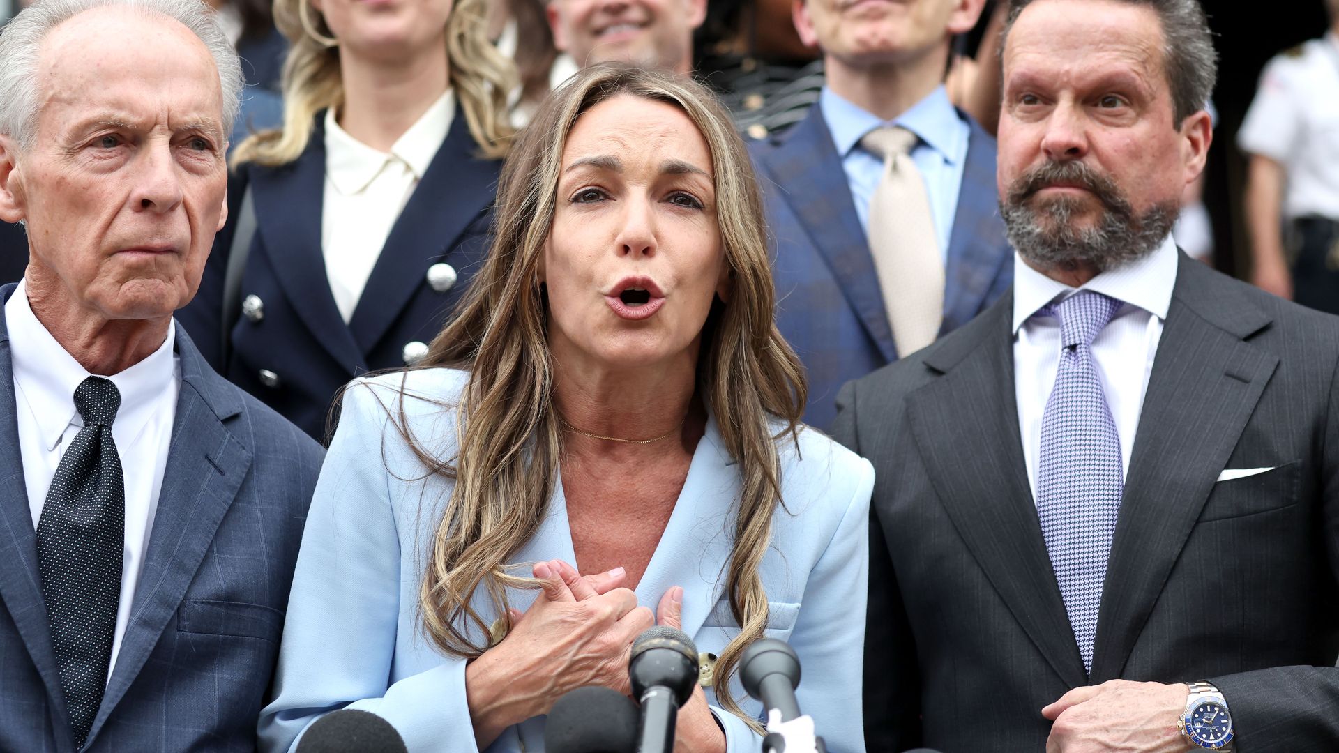 A woman in a light blue blazer speaking passionately at microphones, flanked by men and women in formal attire, standing in front of an audience.