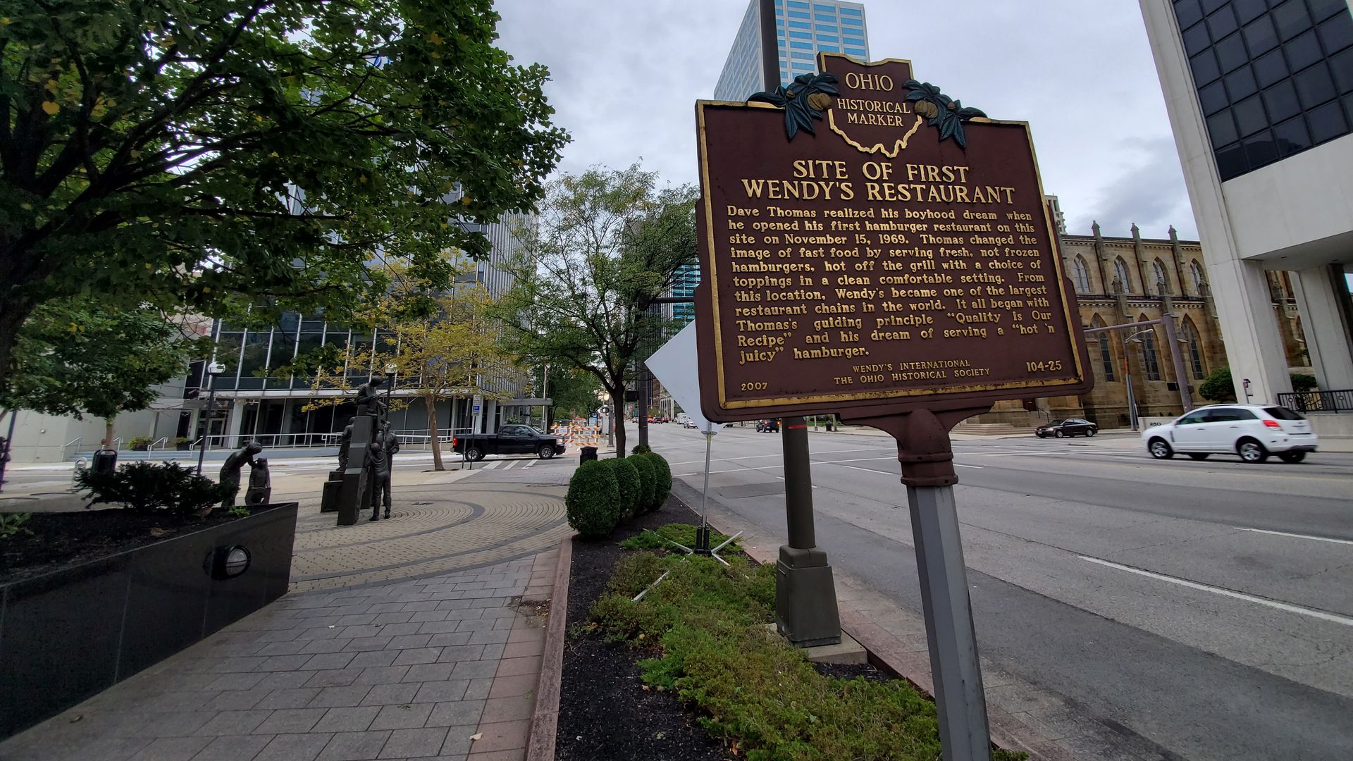 A historical marker rests on a pole in between a sidewalk and a Columbus street. 