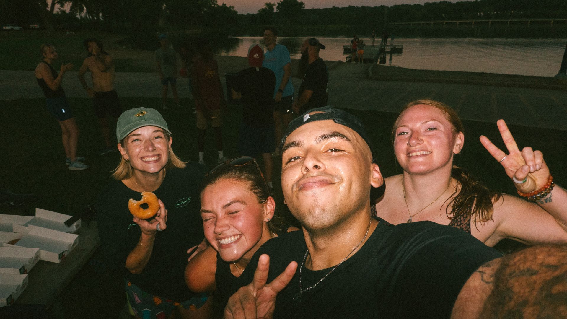 Group selfie at dusk by a lake. Foreground shows three friends smiling; man center holds camera, woman right peace-signs, woman left eats a donut. Background people, trees, and a pier over water.