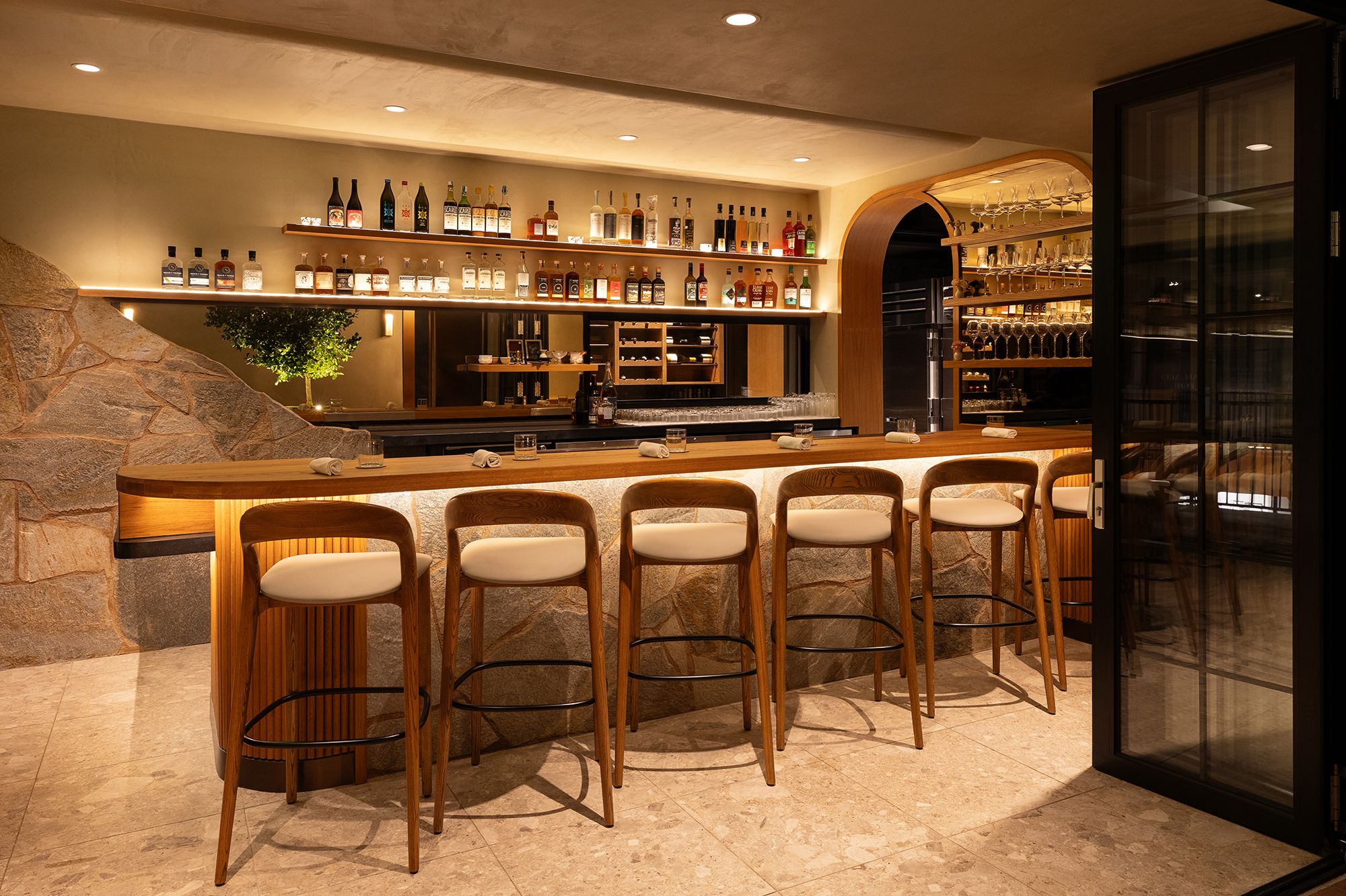 A stylish restaurant interior with warm amber lighting, curved wooden arch, and a long wooden counter with high-top chairs. Bottles line the back shelves, glassware on racks, stone wall left, tiled floor.