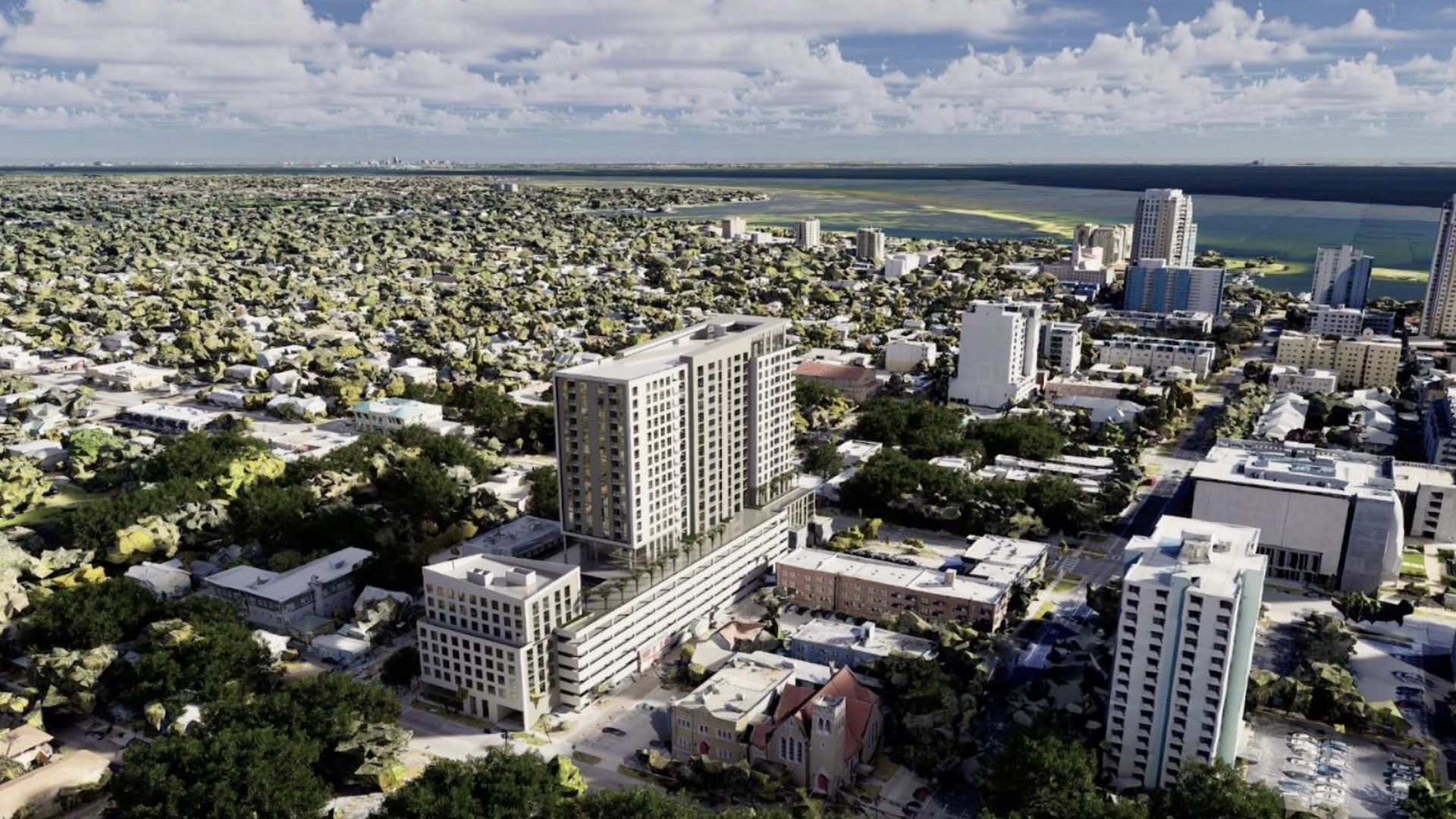 An aerial view of a coastal city: dense residential blocks with green trees, a cluster of tall modern apartment towers near the shore, and a bright blue sky with white clouds.