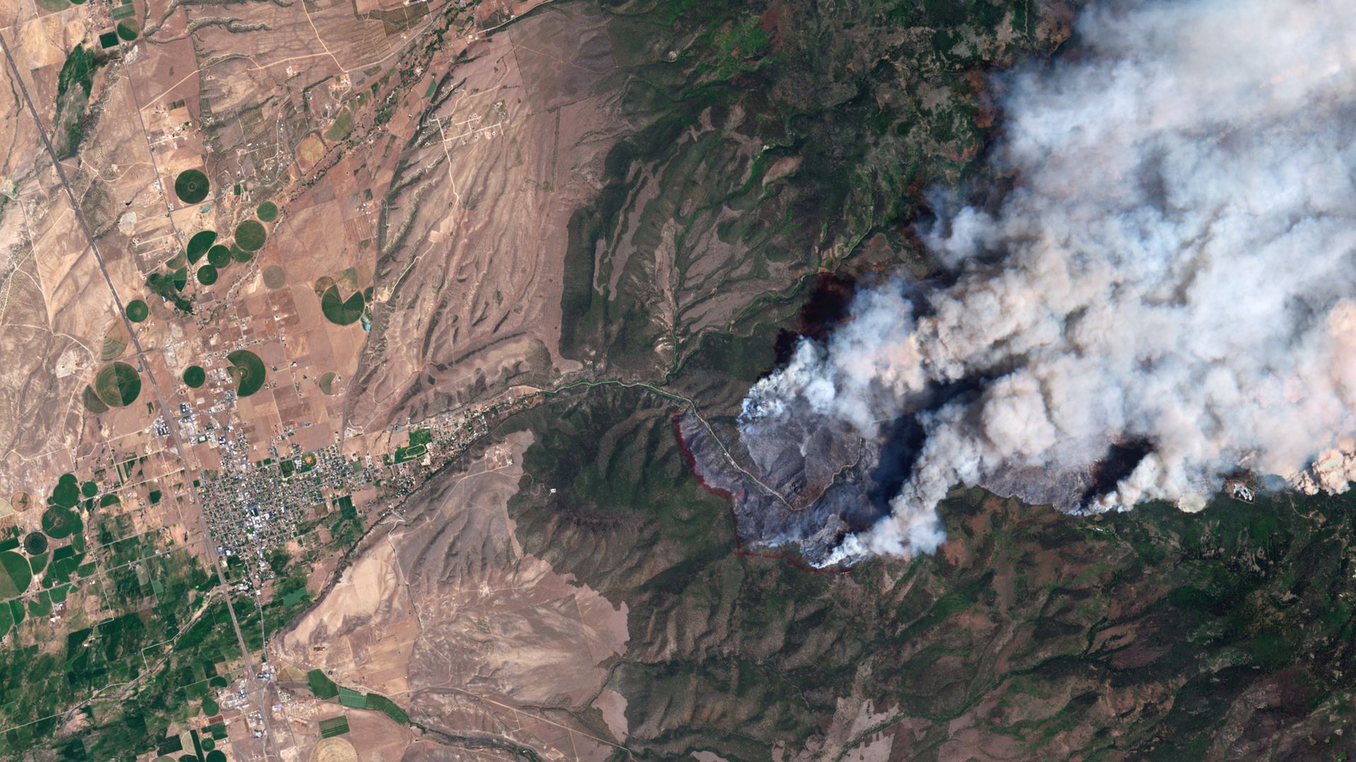 Satellite image showing arid farmlands with numerous green circular center-pivot fields on the left, a hillside valley in the center, and a broad wildfire smoke plume billowing across the right.