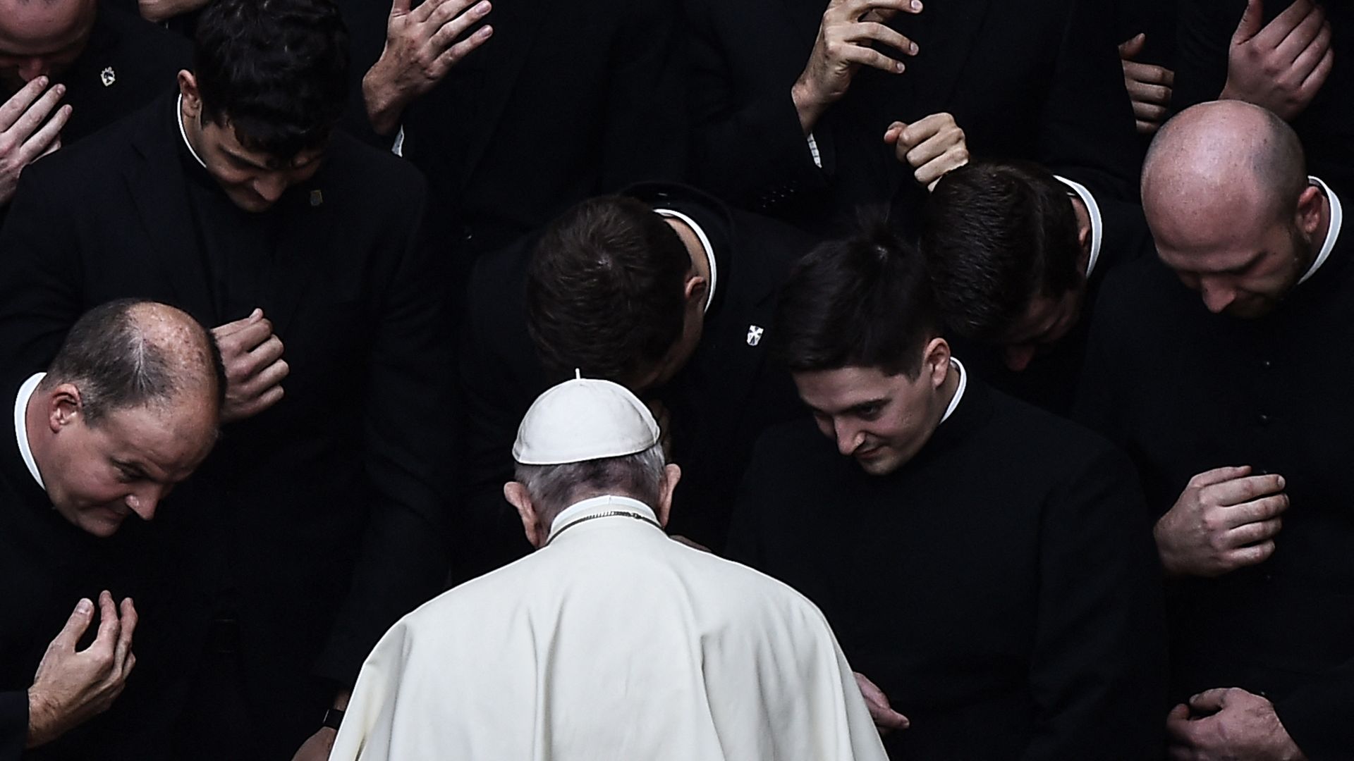 Pope Francis is viewed, in white, from behind, as clergymen, in black, bow toward him in prayer.