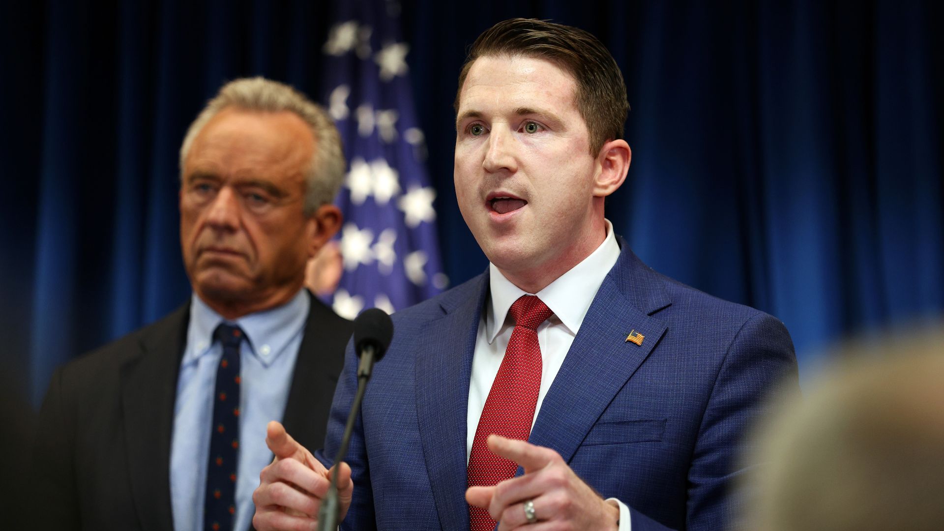 Colin McDonald in a blue suit and red tie speaks at a press conference, gesturing with his hands; Robert F. Kennedy stands behind him, with an American flag and a dark blue backdrop.