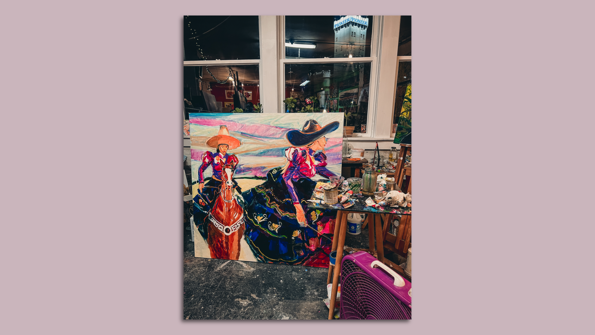 A painting of two women on horses sits in a working studio with a tower building seen through the window.