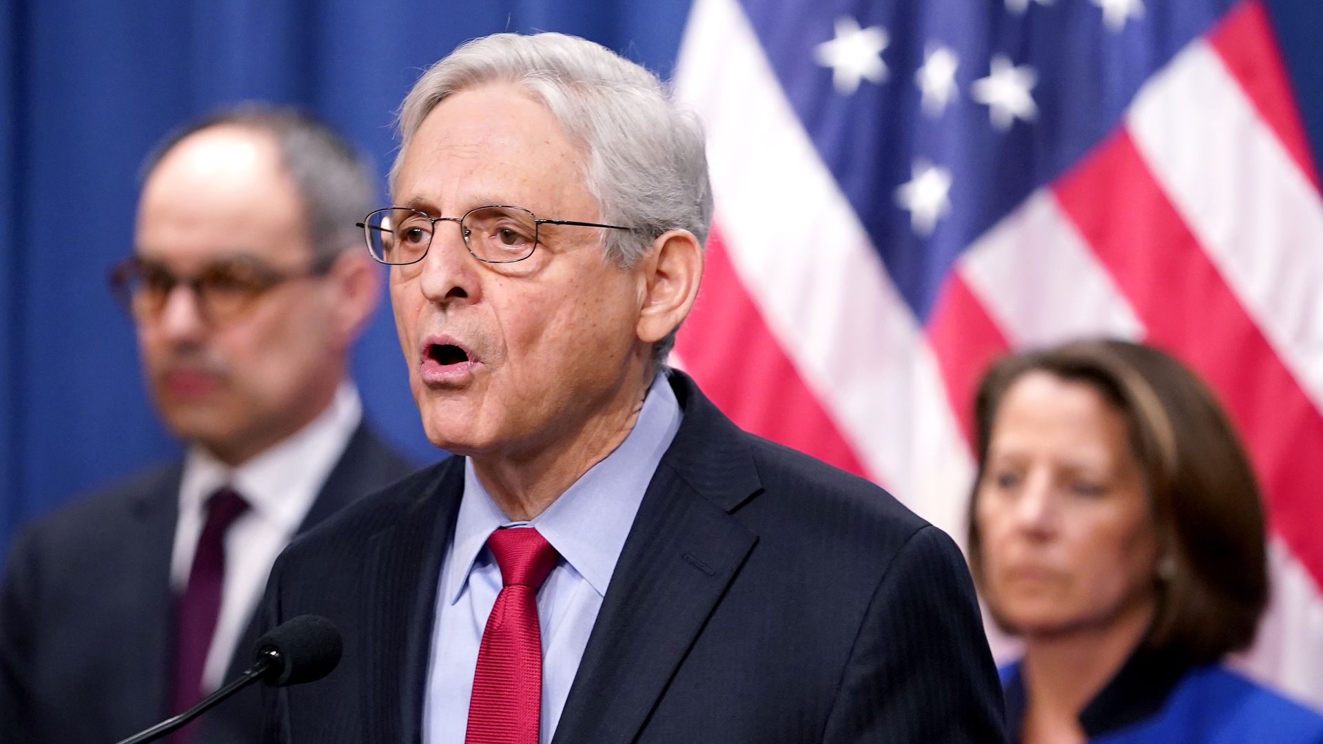 Attorney General Merrick Garland, wearing a dark blue suit, light blue shirt and red tie, flanked by officials standing against a blue curtain and an American flag.