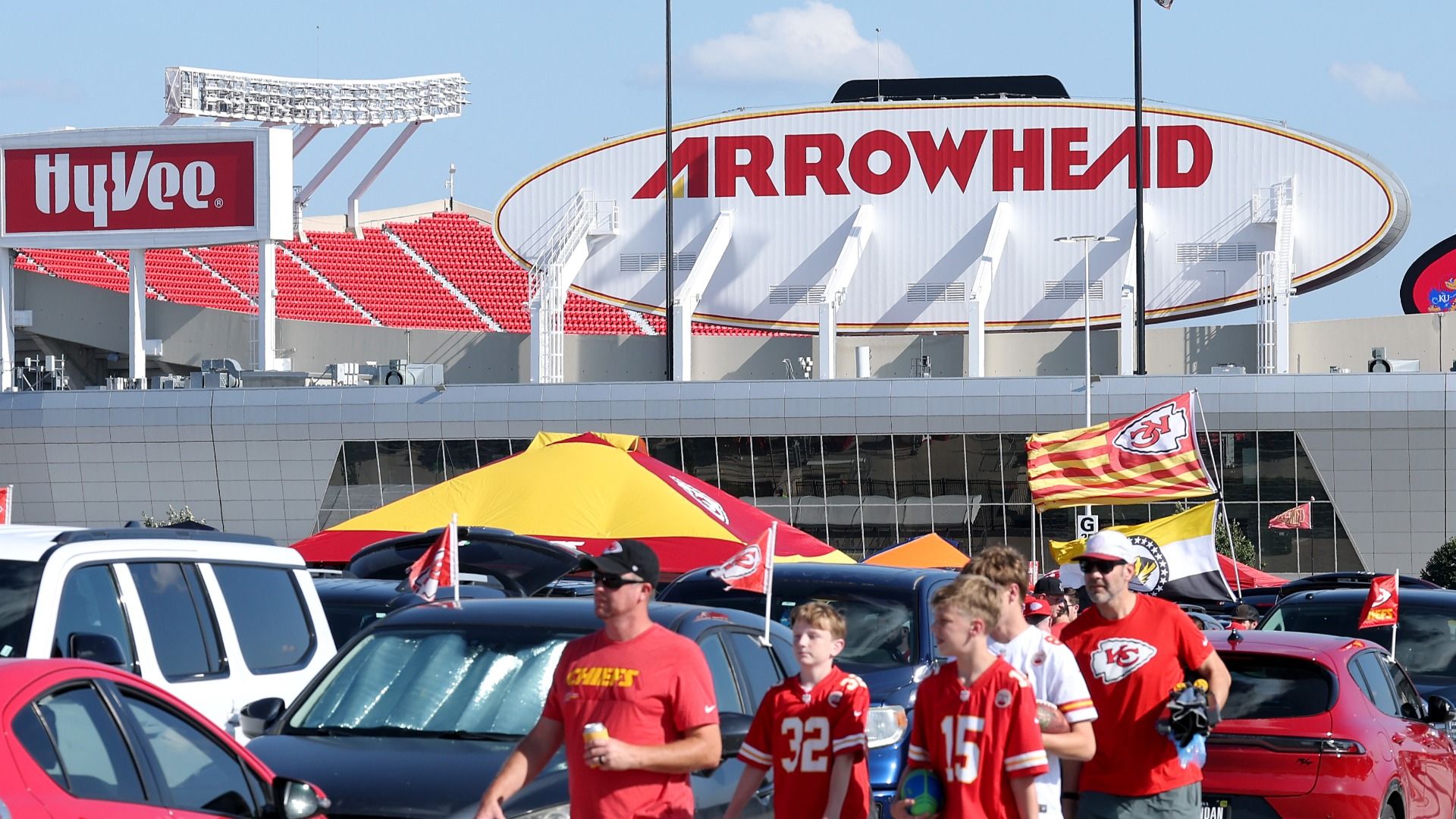 Group of young and old fans in Chiefs shirts, one in a black hat carrying a drink, walk outside of the Chiefs stadium with signs in the background saying "ARROWHEAD" and "HYVEE."