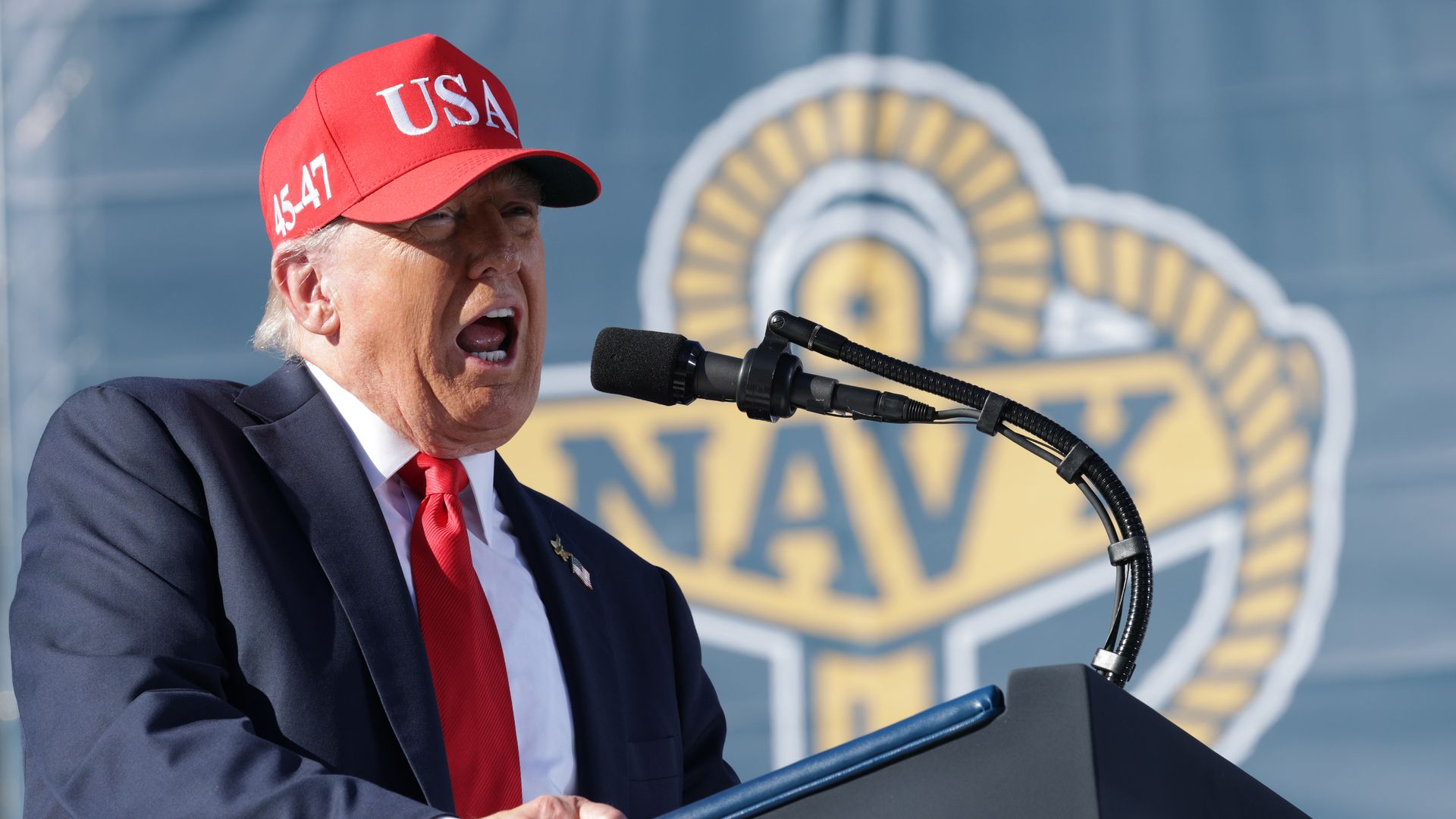 President Trump, wearing a red hat with white "USA" writing, a navy jacket with a US flag, white shirt and red tie, makes remarks into a mic in front of a sign saying "Navy."