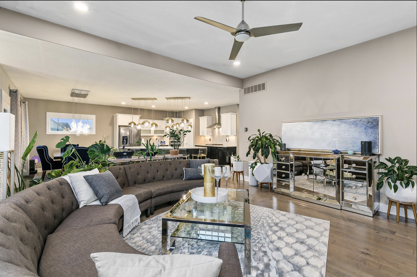 Modern open living room and kitchen with large brown curved sofa, glass coffee table, several green plants, mirrored sideboard, white and gray accents, and a ceiling fan.