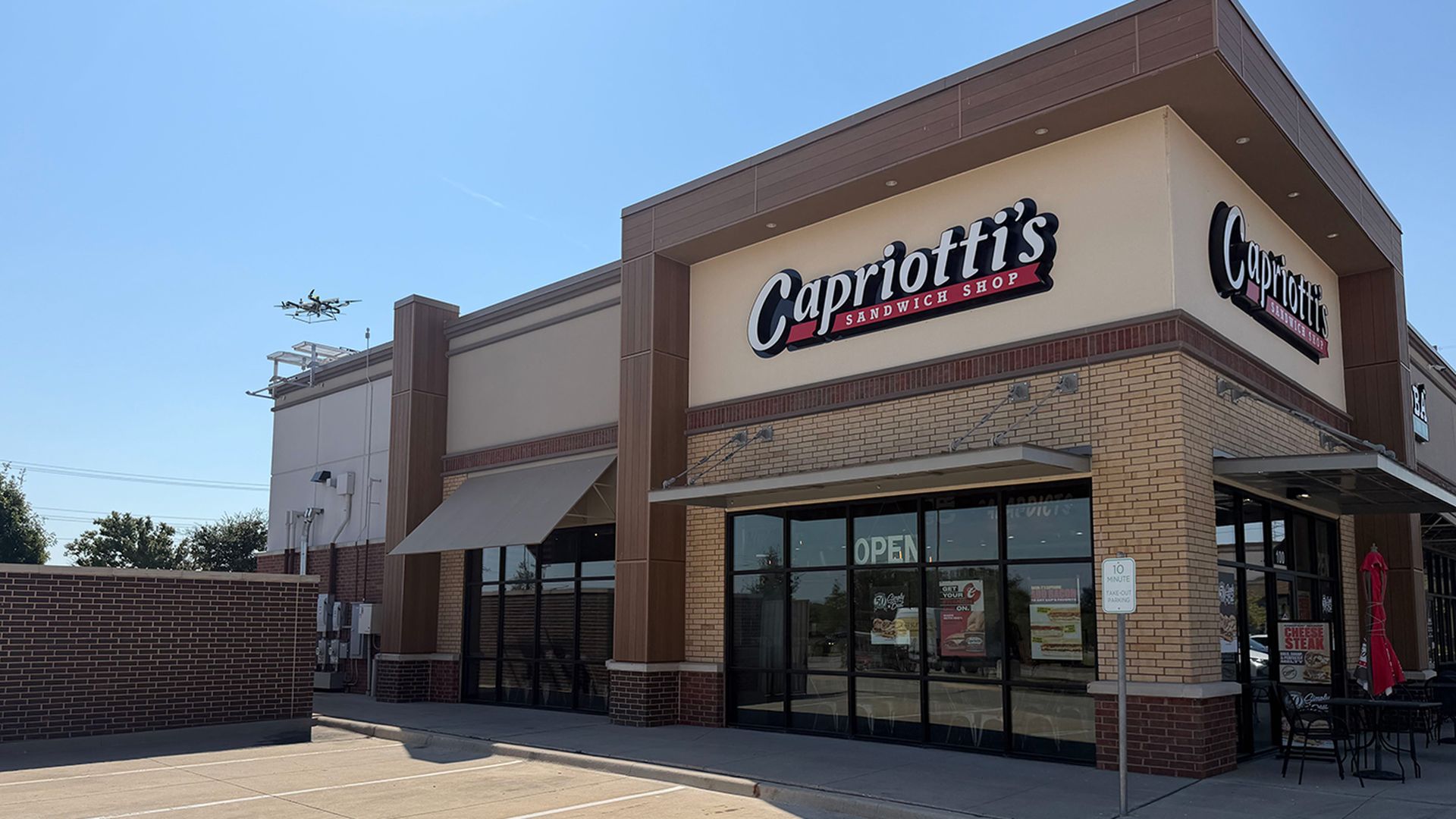 Exterior of Capriotti's Sandwich Shop in a tan brick building with brown trim, a large sign and glass storefront. On the roof, to the rear of the building is a drone dock, with a delivery drone hovering over it.