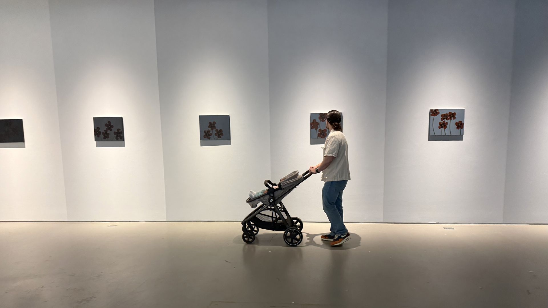 A person in a light shirt and jeans pushes a baby stroller through a white-walled art gallery, pausing before small square blue-gray canvases of red flowers.