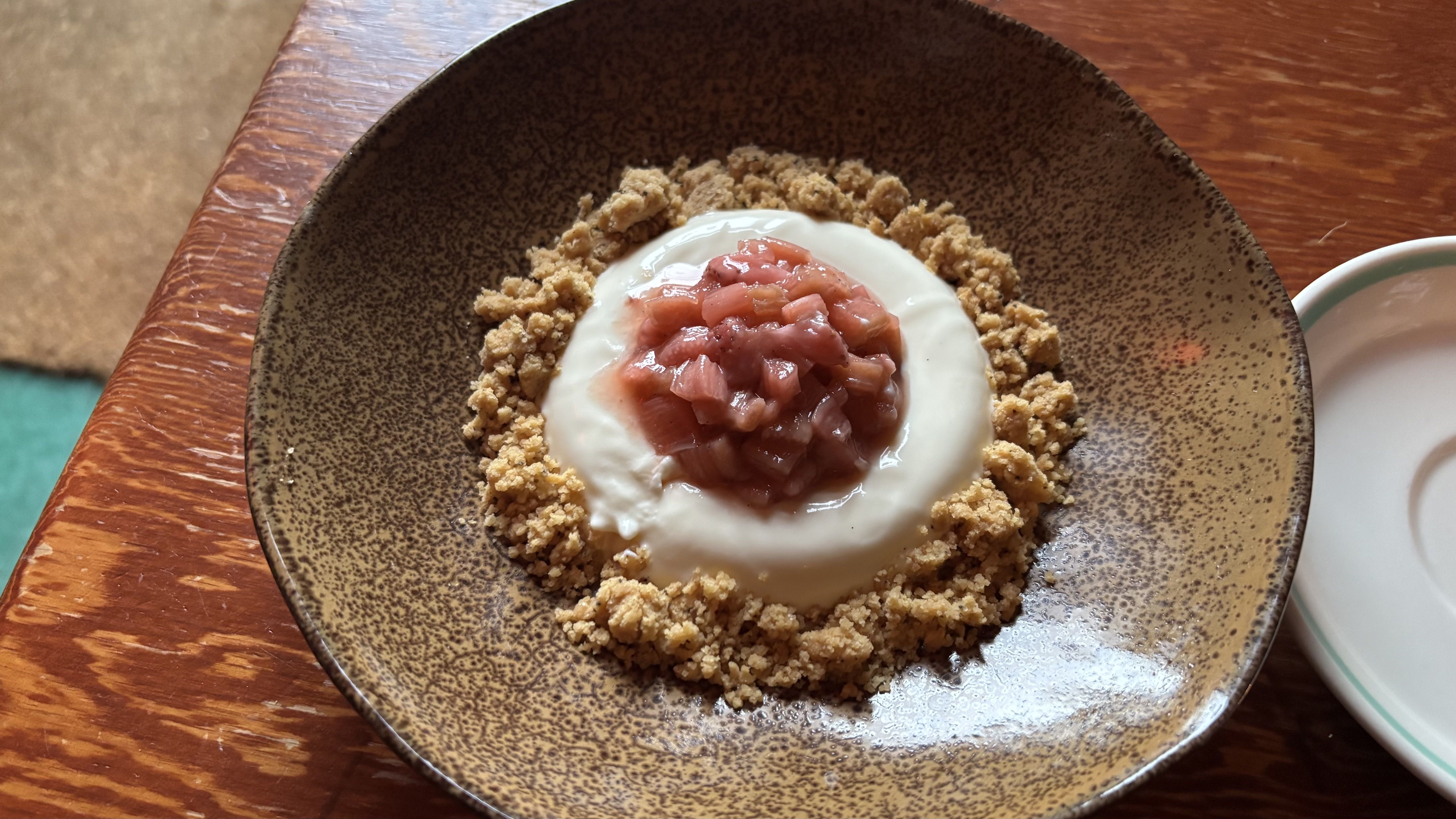 Brown speckled bowl on a wooden table holds a dessert with a white cream center topped with pink diced fruit, surrounded by a ring of crumbly granola crumble.