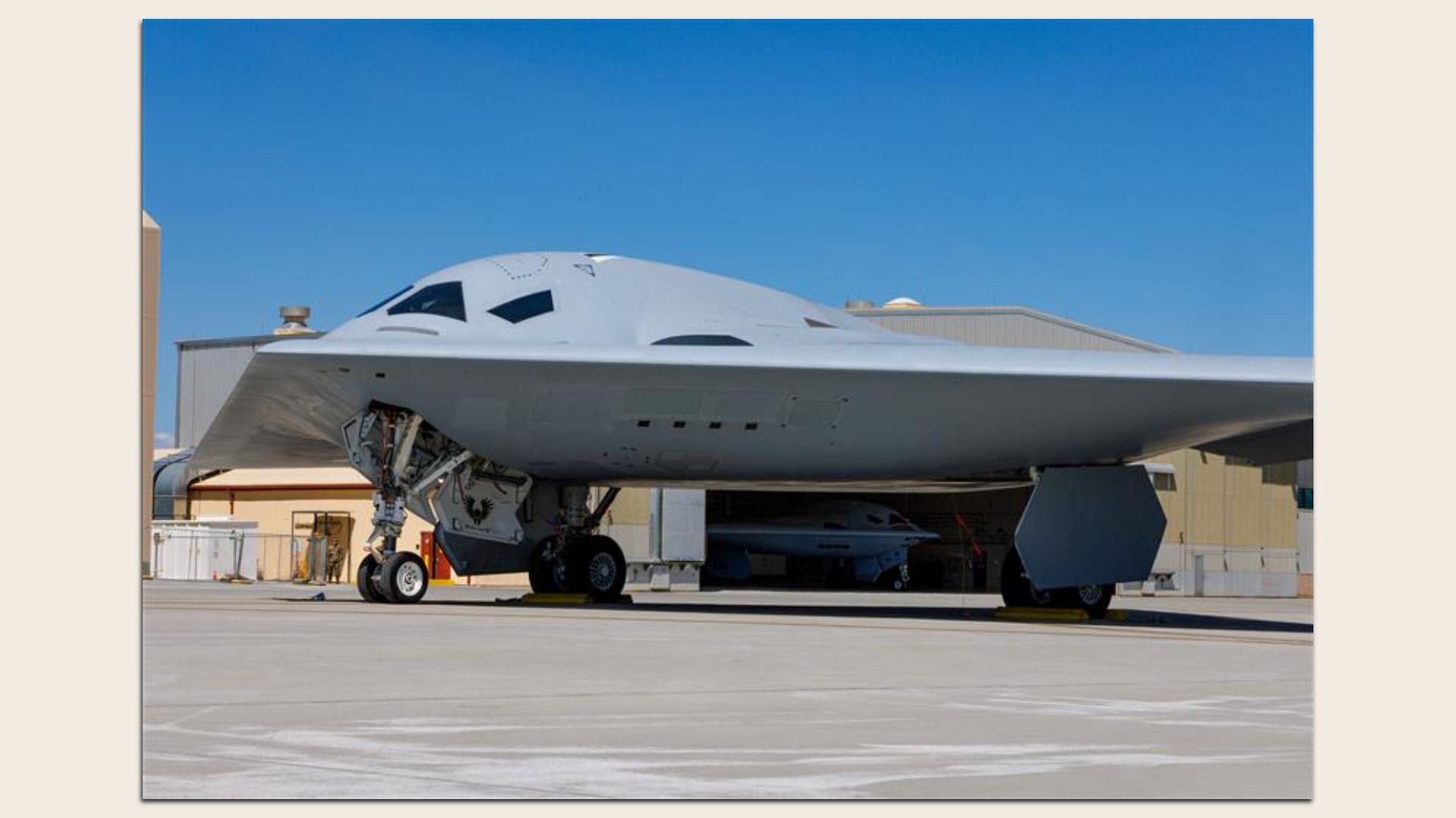 Two B-21s, futuristic bombers, are seen. One is outdoors on the tarmac. The other is inside a hangar. The sky is clear and blue.