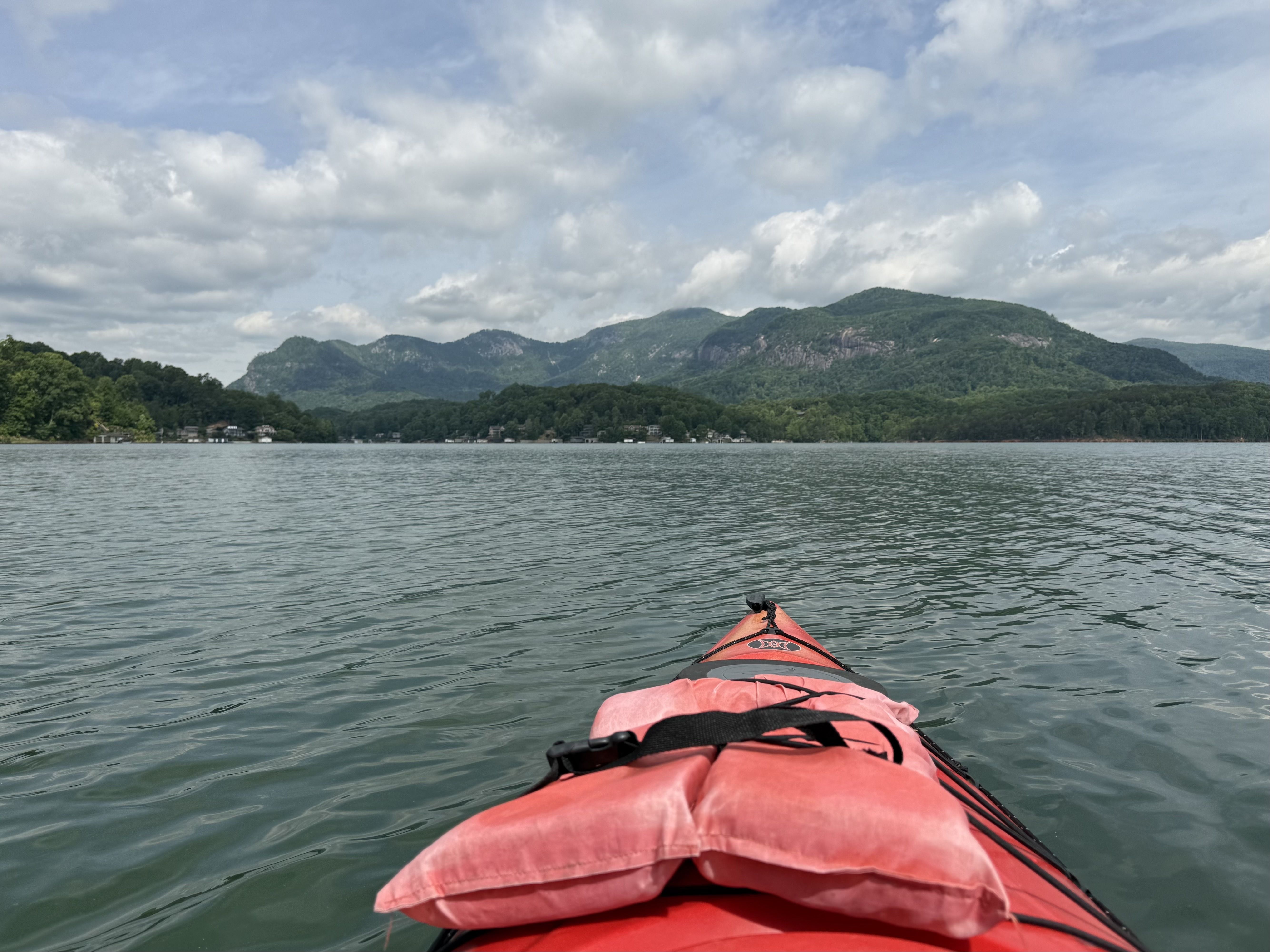 First-person view from a red kayak on a calm lake; orange life jackets visible, with forested hills and a cloudy blue sky in the distance.