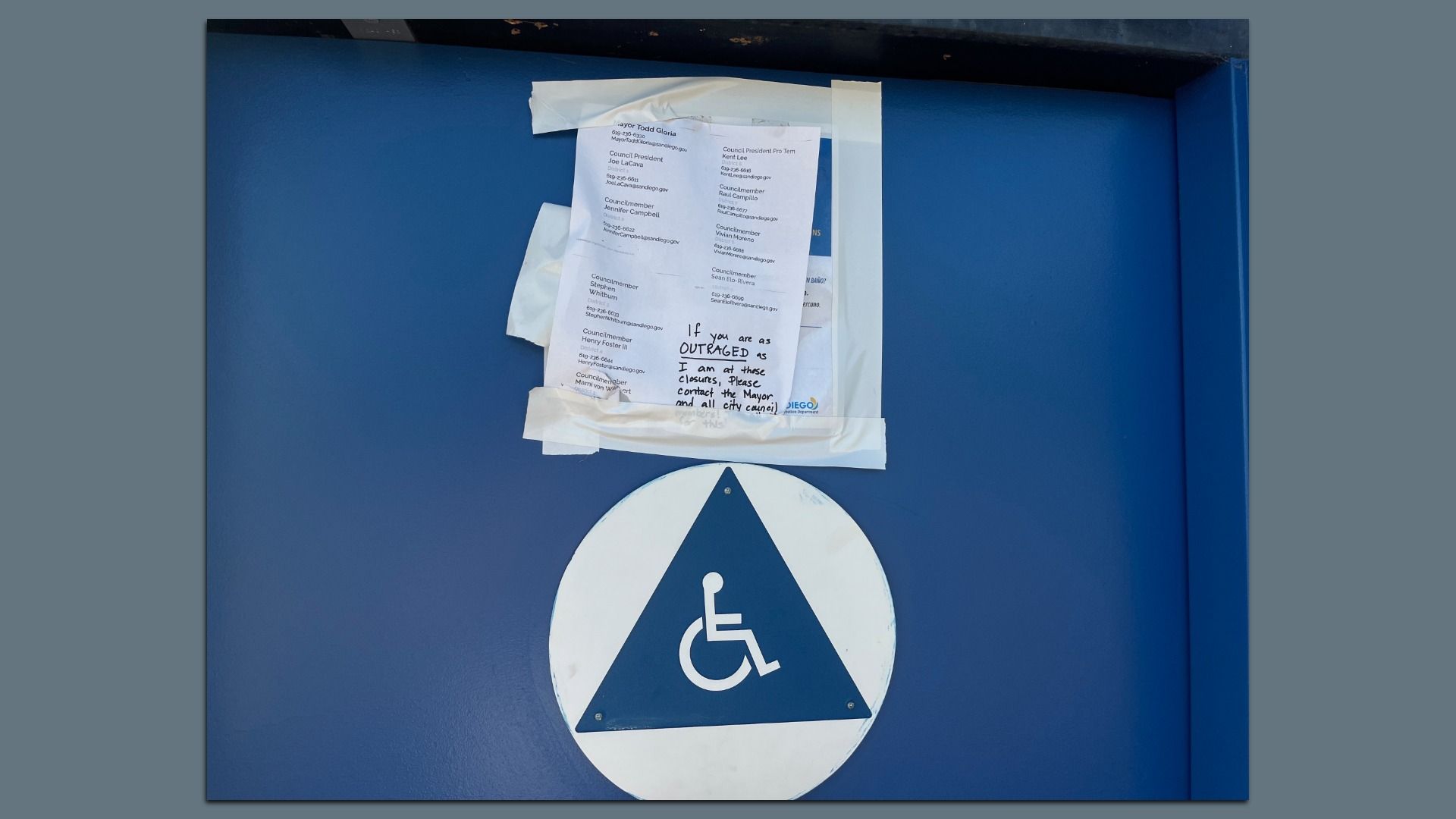 Blue wall with a large handicap-accessibility sign at the bottom. A stack of white posters taped to the wall, including a handwritten note about being outraged and contacting the mayor and council.