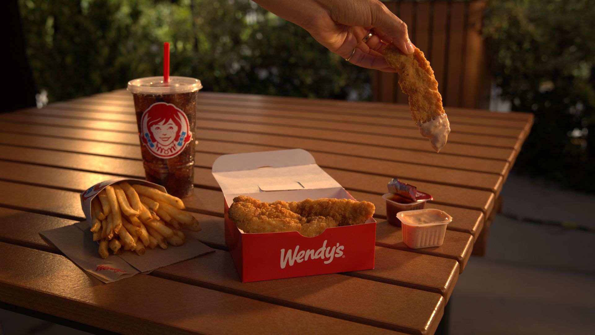 Hand dipping a crispy chicken strip into sauce at a wooden outdoor table with Wendy's fries, a clear cup filled with soda with a drawing of the Wendy's mascot and a red Wendy's box containing more chicken strips.
