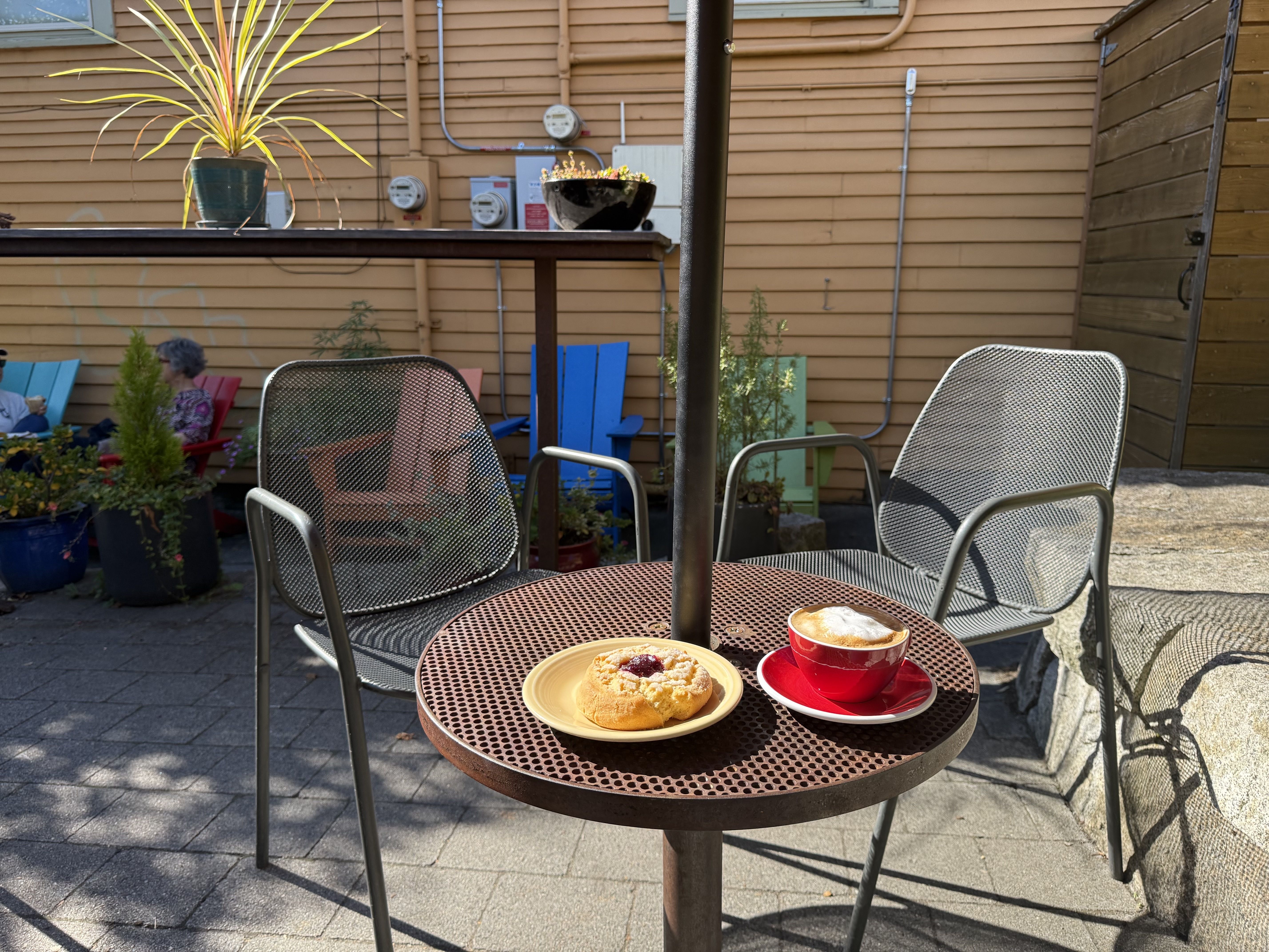 Outdoor café table with metal mesh chairs, a coffee cup in a red saucer, and a pastry on a beige plate, next to a tan wood siding wall and colorful Adirondack chairs in background.