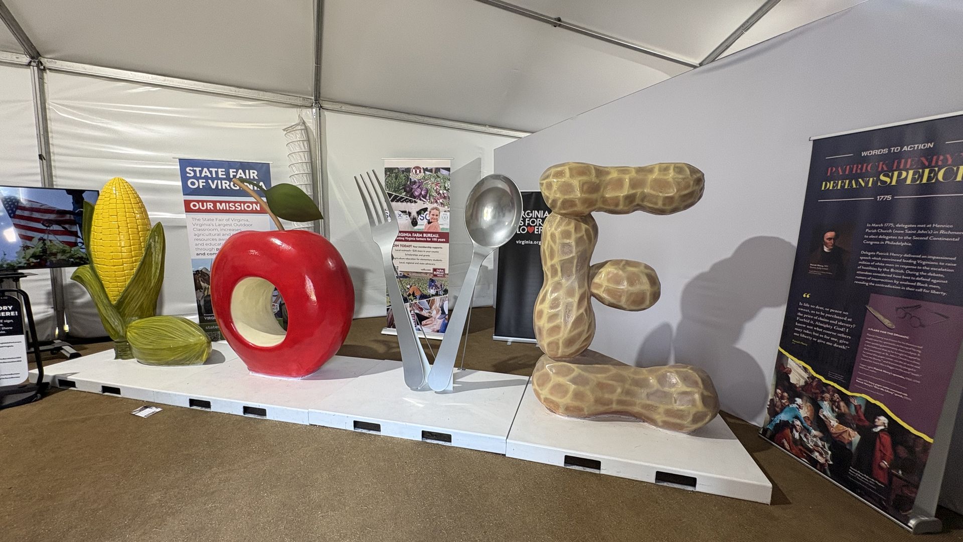 Exhibit booth inside a white tent with giant props: a yellow corn on the cob, a red hollow ring, oversized peanut sculptures, and big forks. Banners read "State Fair of Virginia" and "Patrick Henry" poster.