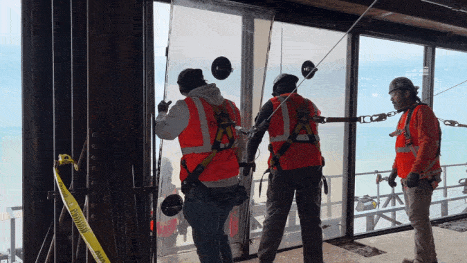 Several construction workers in orange safety vests with harnesses stand along a glass wall on a high-rise, installing panels; yellow caution tape and a blue ocean view are visible outside.
