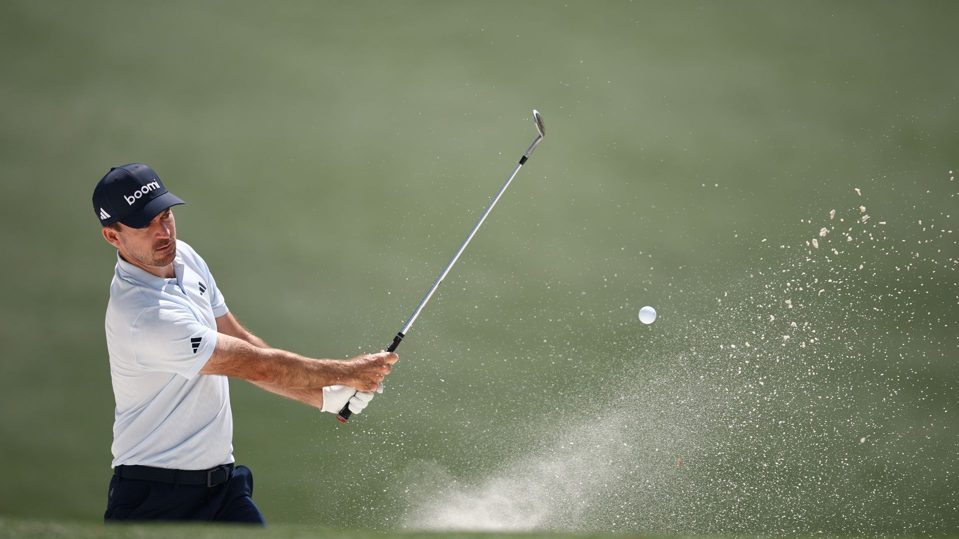 A golfer in a white shirt and dark baseball cap in mid swing with the ball in the air. 