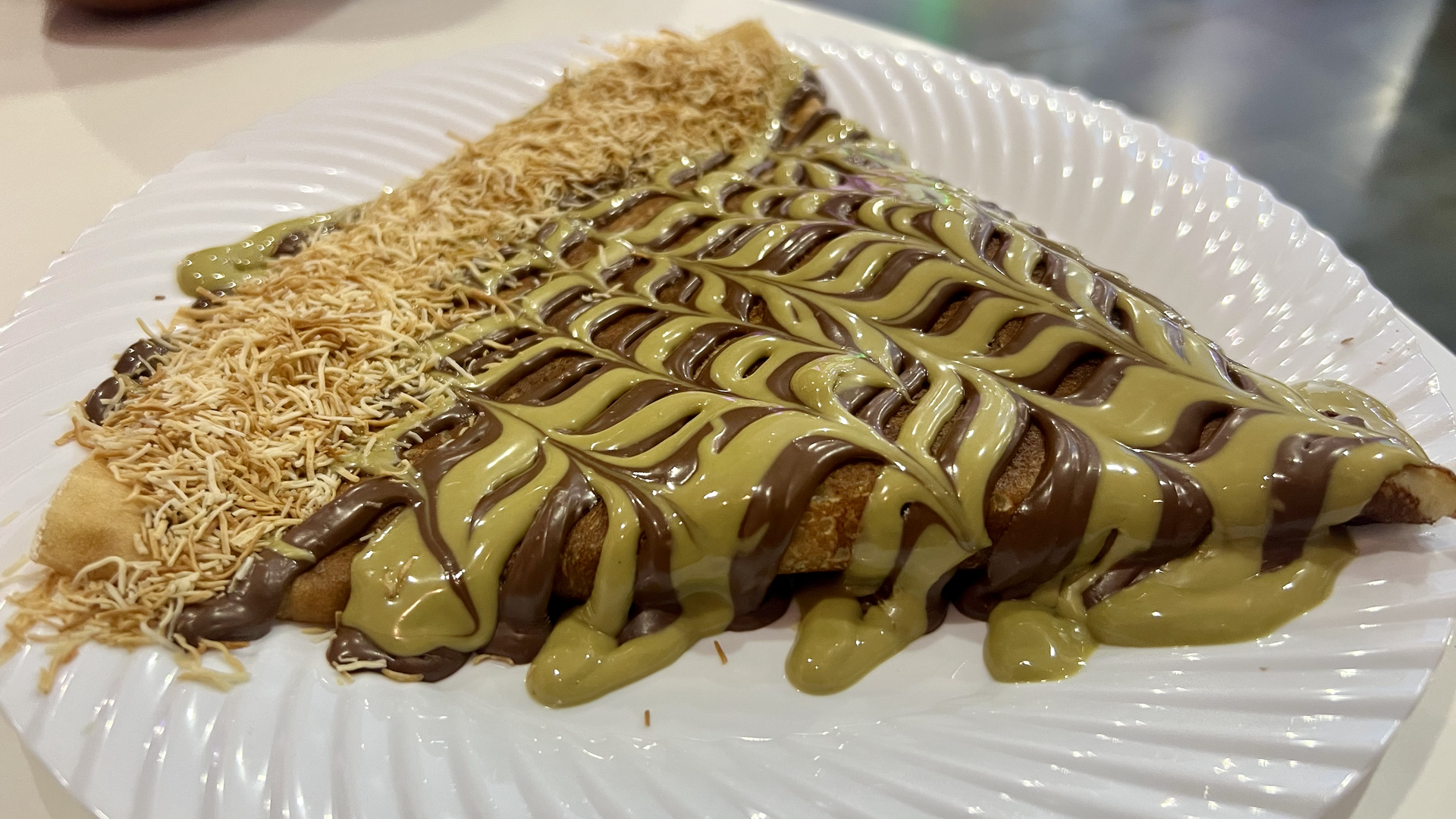 Close-up of a wrap sandwich cut in half, showing pink meat, pickles, tomatoes, red onions, and herbs inside a toasted flatbread, with a sauce cup and chips in the background.