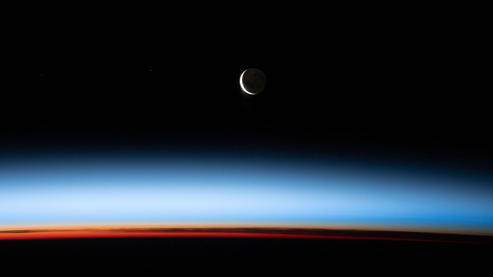 A crescent moon shines above the limb of the Earth with the blue glow of the atmosphere 