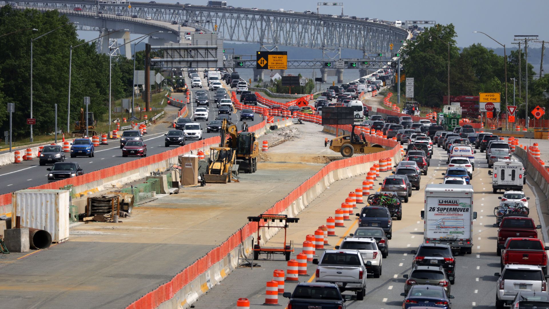 Busy highway under construction with orange barriers and traffic cones, construction equipment in the foreground, heavy cars on both sides, and a large steel bridge spanning the distance.