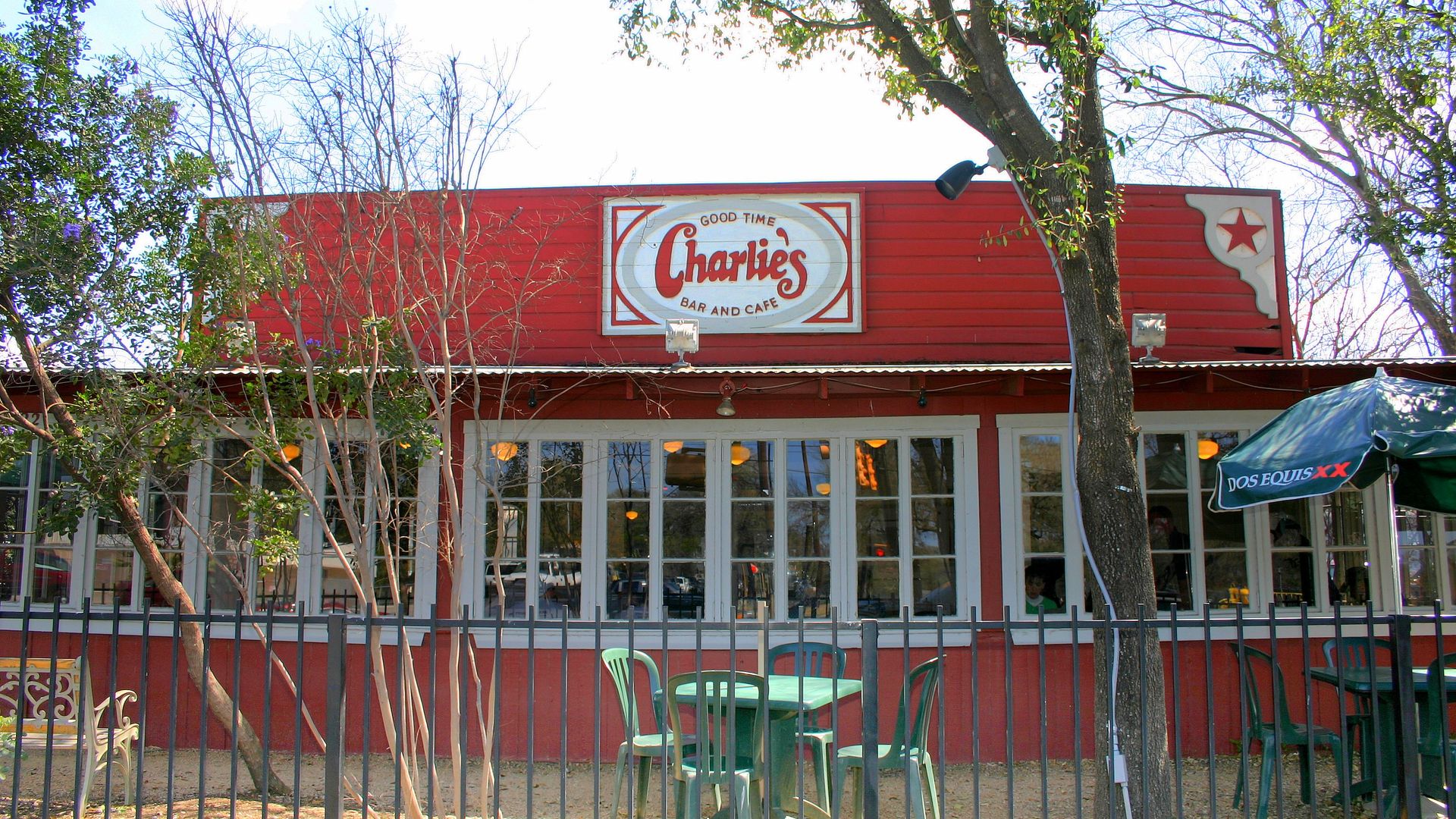 Exterior of a red building labeled "Charlie’s Bar and Cafe" with outdoor green tables and chairs, a metal fence, bare trees, and a Dos Equis umbrella.