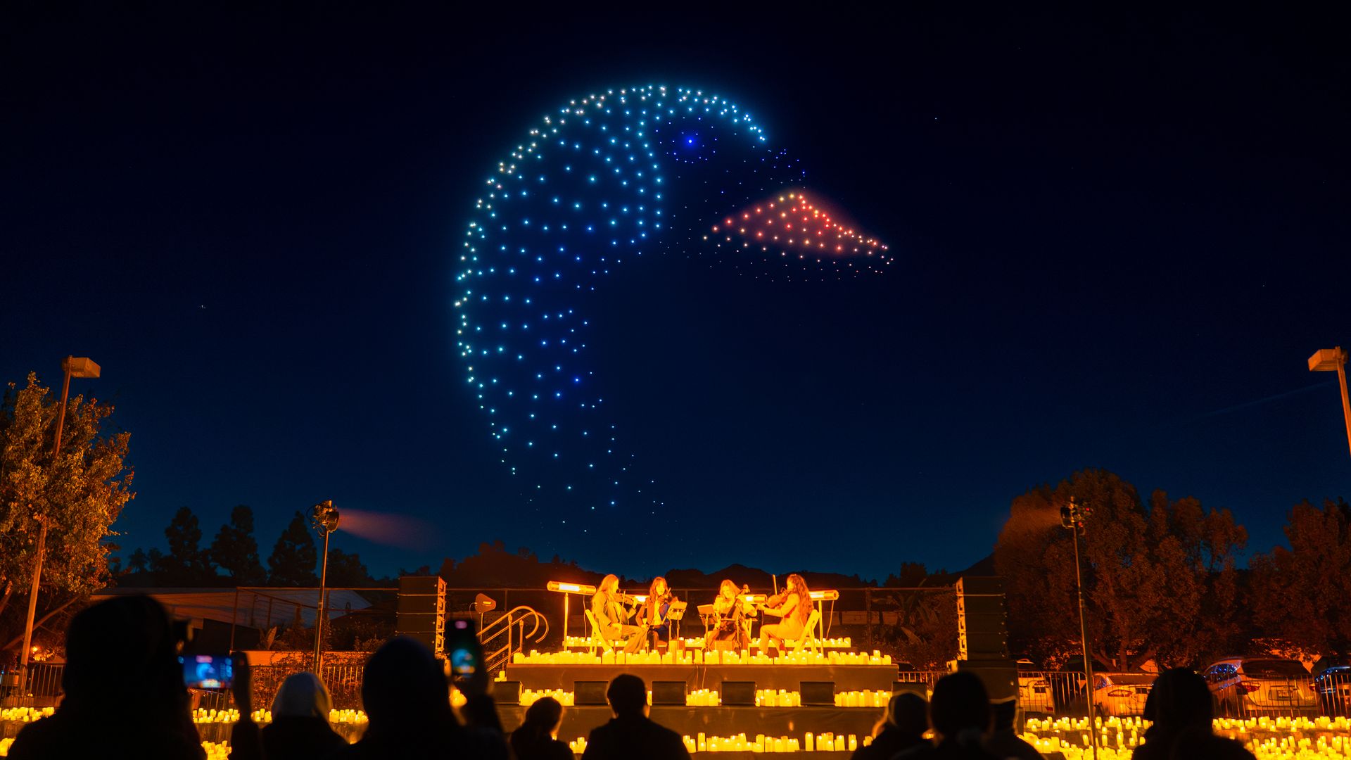 Night scene of a string quartet playing on a candlelit stage outdoors with an audience watching. Above them, blue and orange drone lights form the shape of a duck's head in the dark sky.