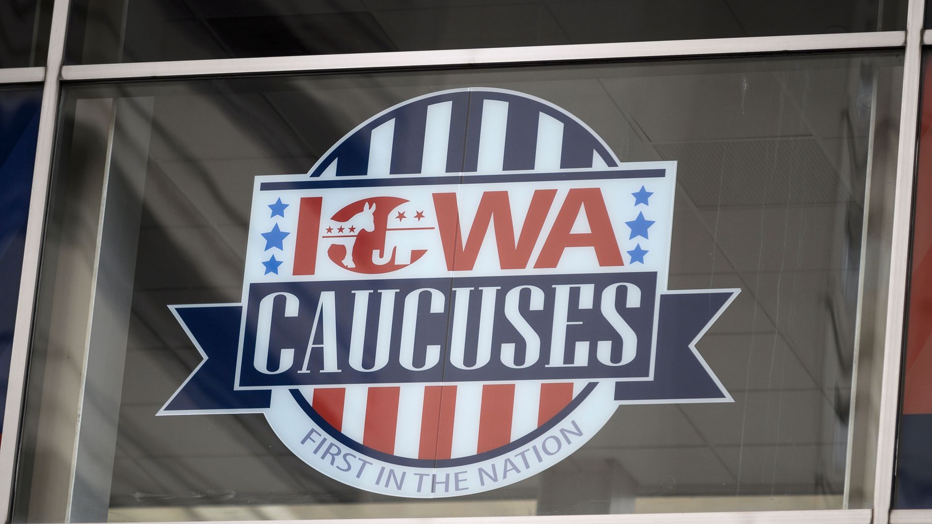 Sign on window for Iowa Caucuses with red, white, and blue colors, featuring a donkey and elephant symbol, stars, stripes, and text "First in the Nation".