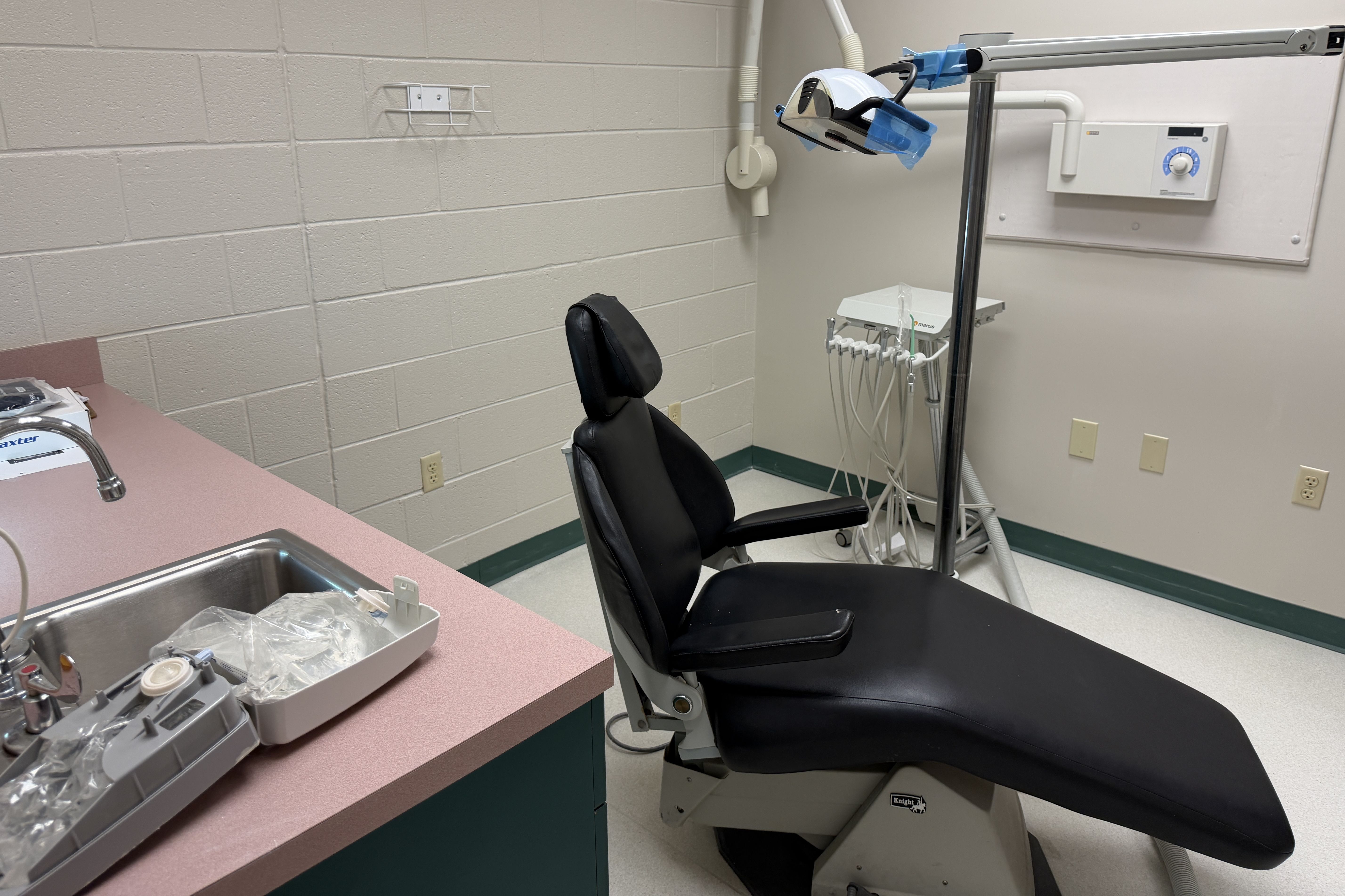 Dental treatment room with a black reclining chair, pink countertop and instrument tray in the foreground, overhead light and dental equipment on a stand, and white cinder-block walls.